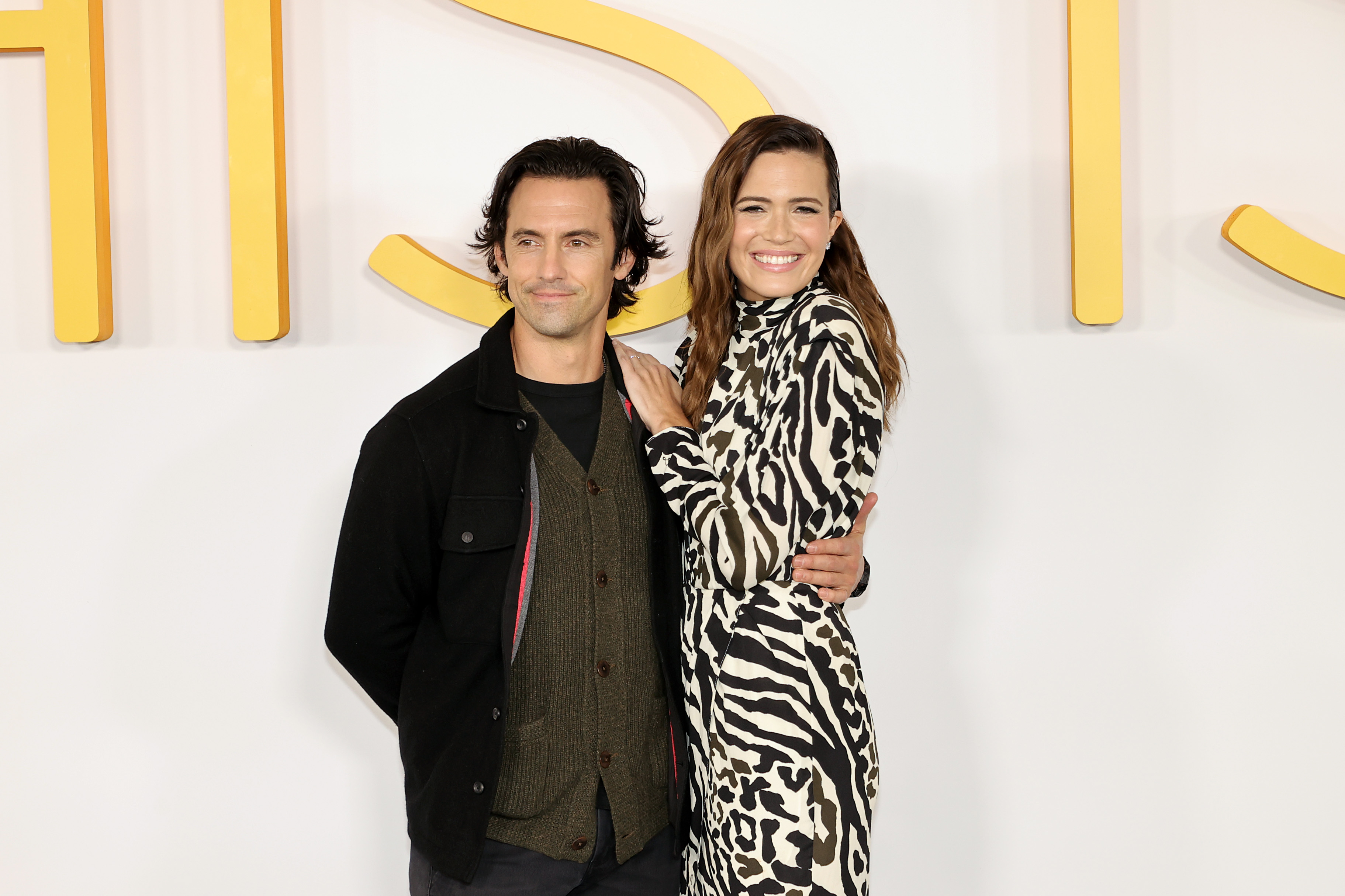 'This Is Us' stars Milo Ventimiglia and Mandy Moore embrace one another as they pose for pictures on a red carpet. Ventimiglia wears a black jacket over a dark green cardigan over a black shirt. Moore wears a zebra-patterned long-sleeved dress.