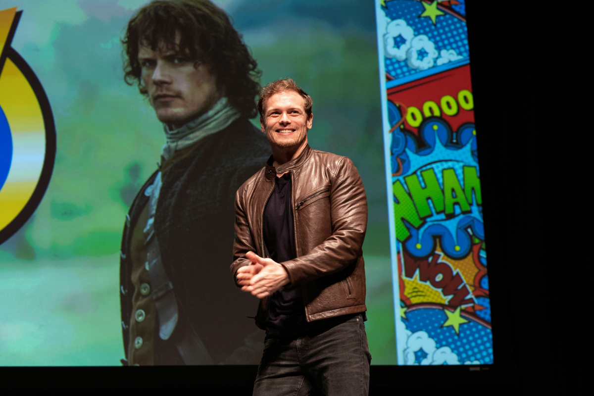 Sam Heughan speaks on stage with a picture of Jamie Fraser behind him during Wizard World Comic Con at Ernest N. Morial Convention Center on January 04, 2020 in New Orleans
