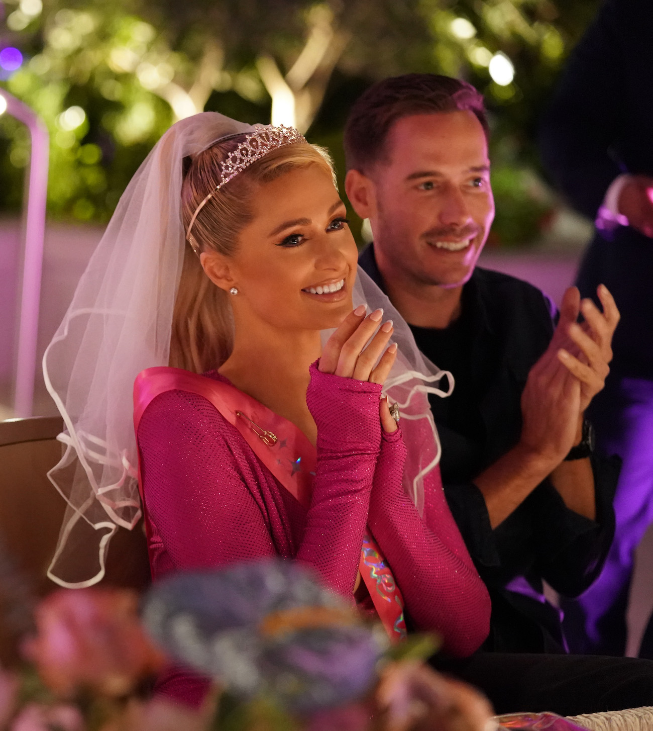 Paris Hilton wears a pink dress and veil as she smiles while sitting next to Carter Reum