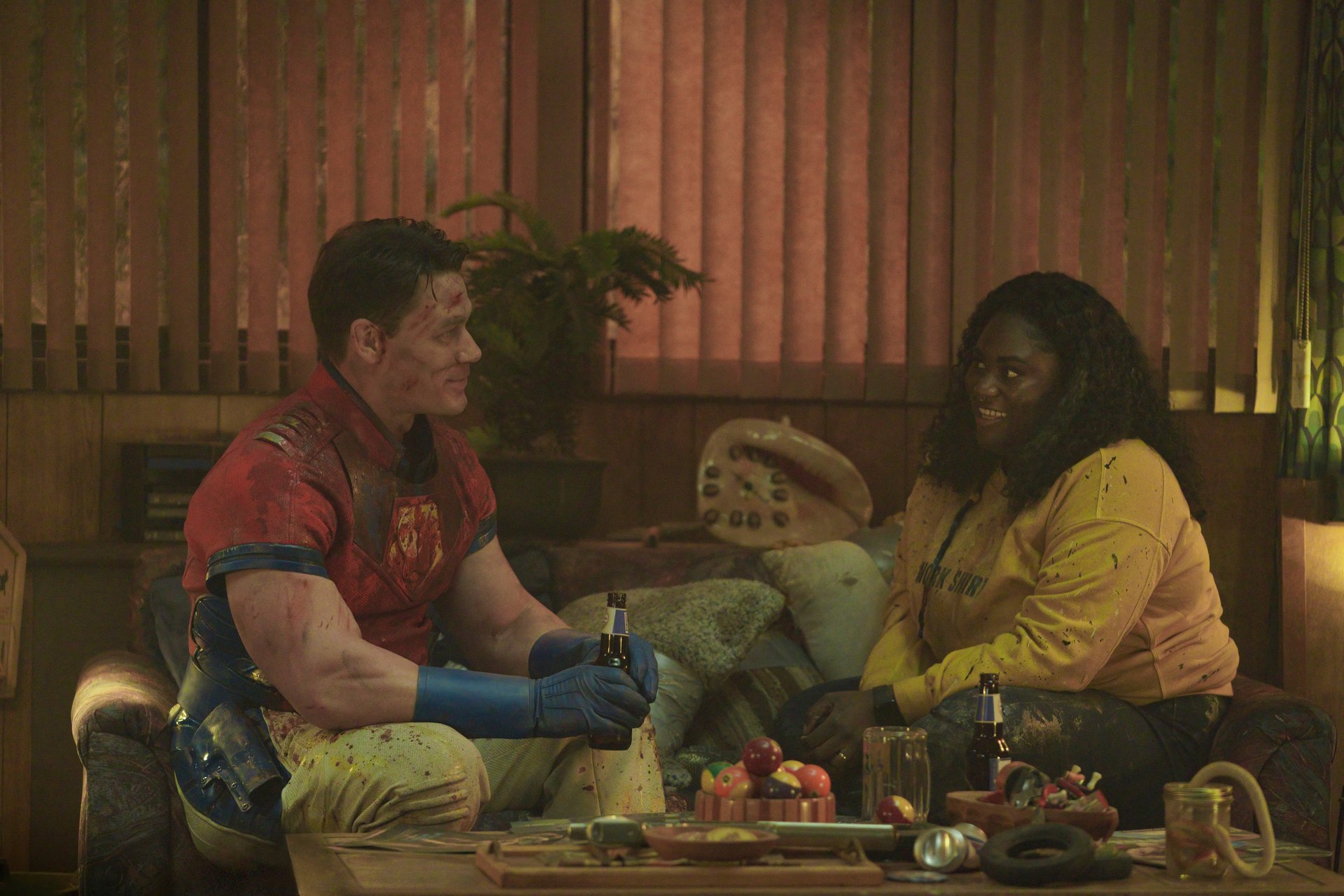 John Cena and Danielle Brooks as Peacemaker and Leota Adebayo in DC's 'Peacemaker' Episode 5. They're sitting on the couch and talking to one another.