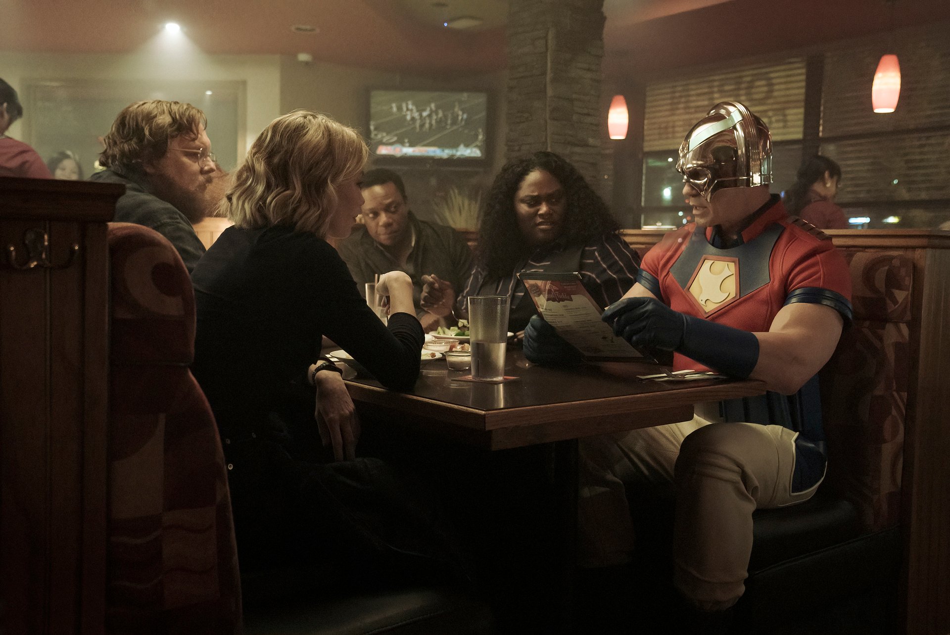 Jennifer Holland, Steve Agee, Chukwudi Iwuji, Danielle Brooks, and John Cena sitting at a restaurant in 'Peacemaker' Episode 1.
