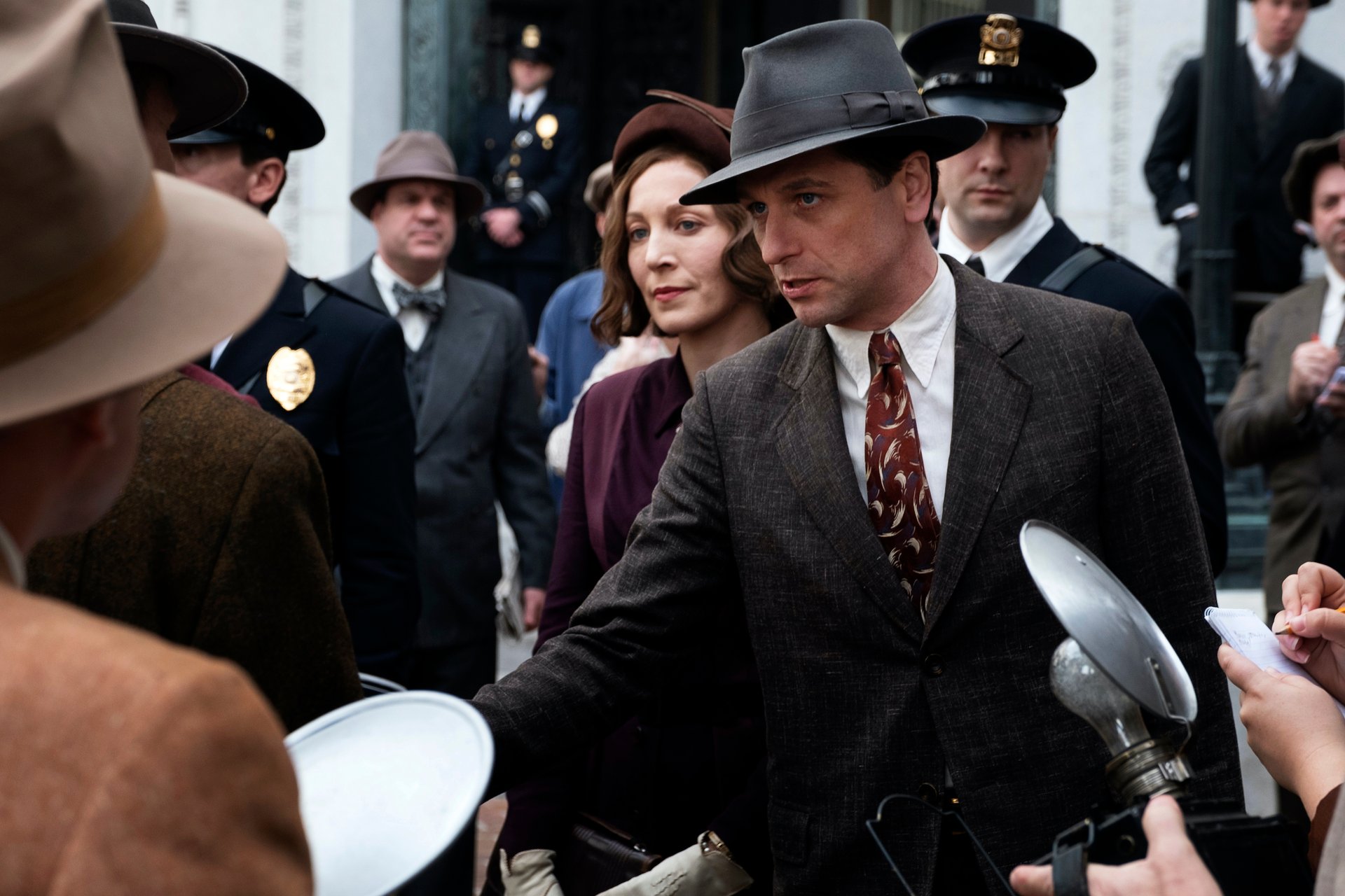 Matthew Rhys as Perry Mason, wearing a hat and talking to reporters holding cameras in an episode of 'Perry Mason'