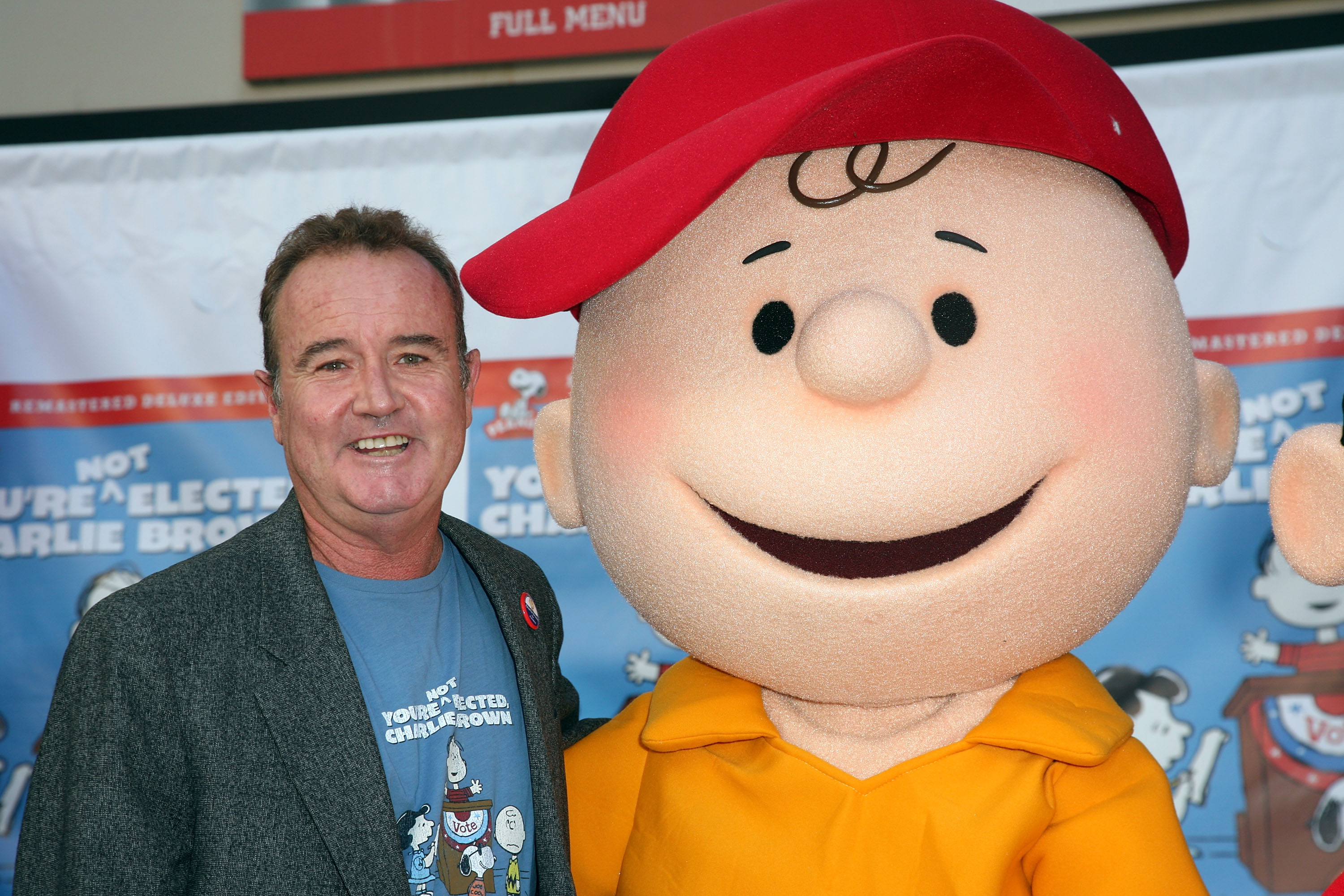 Peter Robbins, wearing a blazer and a Peanuts T-shirt, standing next to a Charlie Brown statue