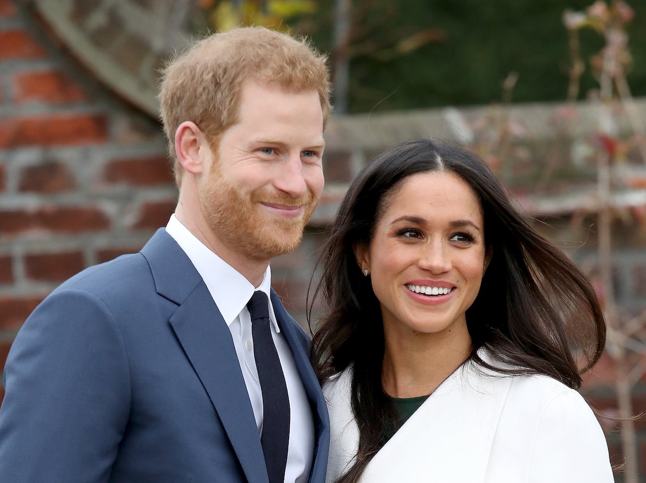 Prince Harry and Meghan Markle looking off-camera and smiling. Harry wears a suit and tie and Meghan is wearing a white overcoat