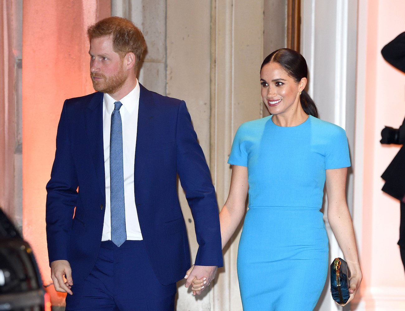 Prince Harry wearing a suit and holding hands with Meghan Markle, who is wearing a blue dress