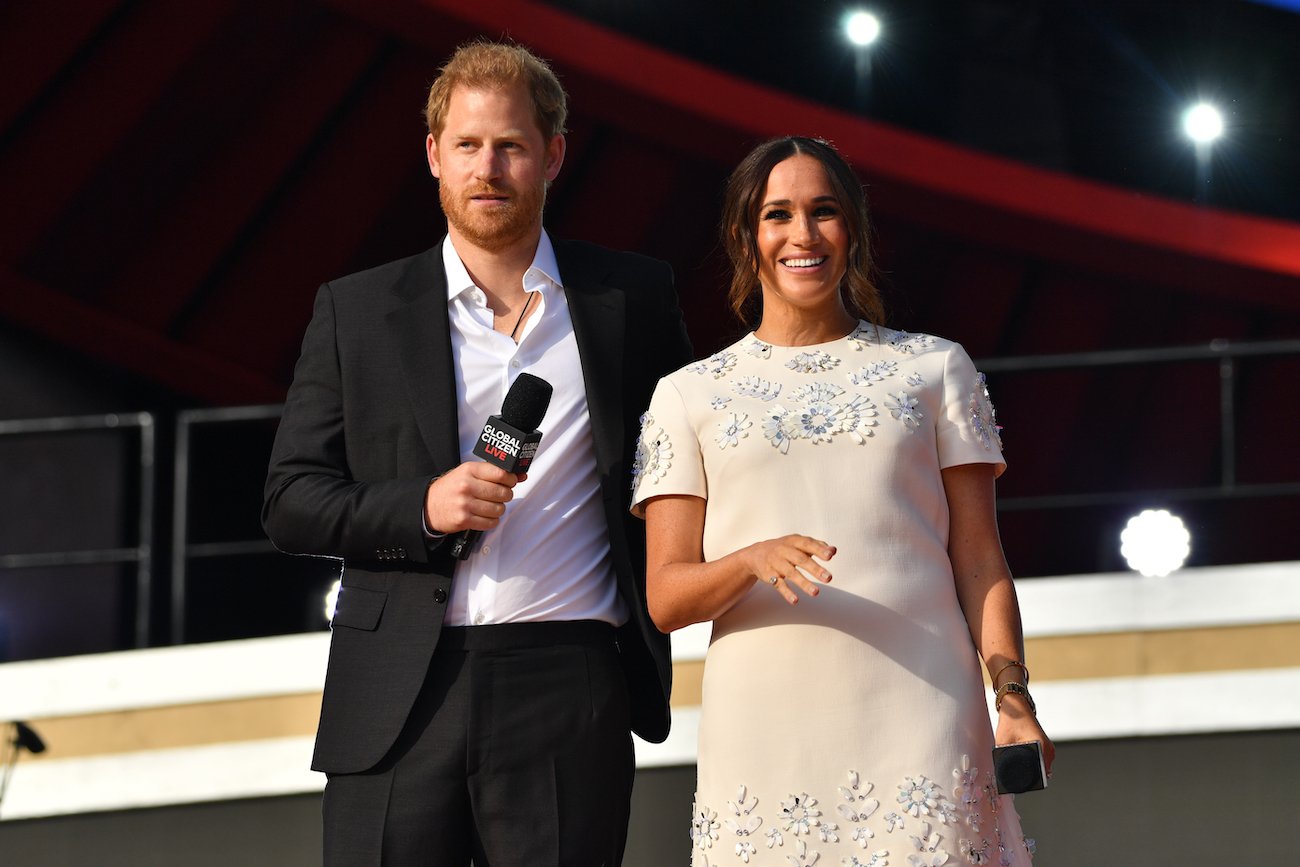 Prince Harry and Meghan Markle look on with Harry wearing a suit and Meghan in a white dress as they stand onstage at Global Citizen Live