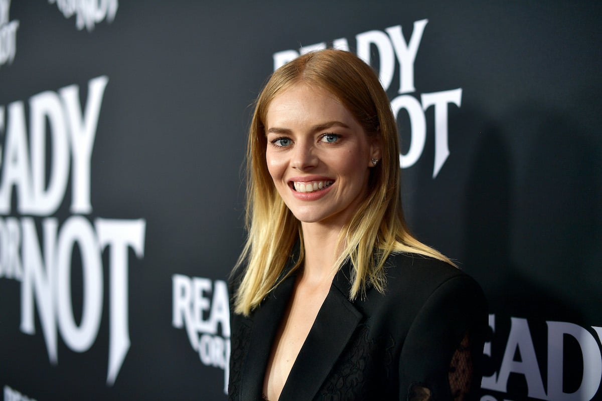 Samara Weaving wears black and smiles in front of the ‘Ready or Not’ logo