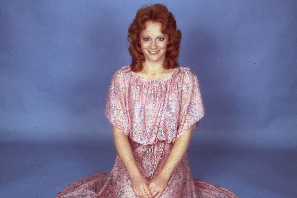 Reba McEntire (c. 1976) sits on her knees in a pink dress spread out around her, smiling with her hands folded on her lap