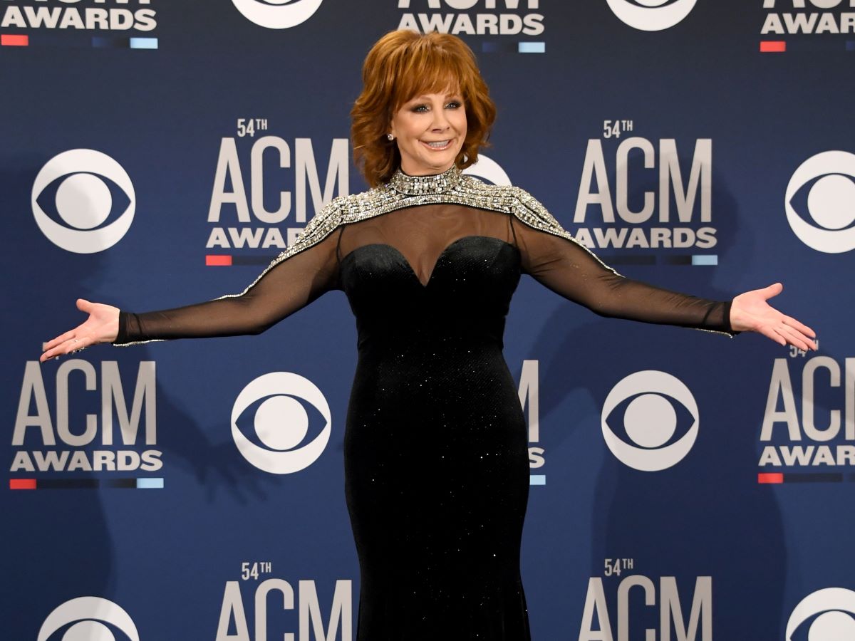 Reba McEntire stands with her hands out to her sides, wearing a black dress with a silver collar and sleeves