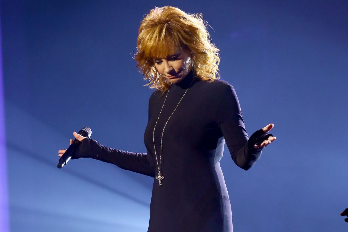 Reba McEntire in a black dress and long cross pendant, holding a microphone in one hand and extending both hands out to the side while looking down.