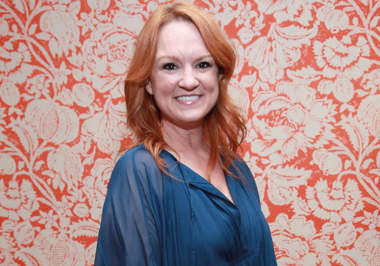 Ree Drummond smiles wearing a blue shirt in front of an orange-printed backdrop