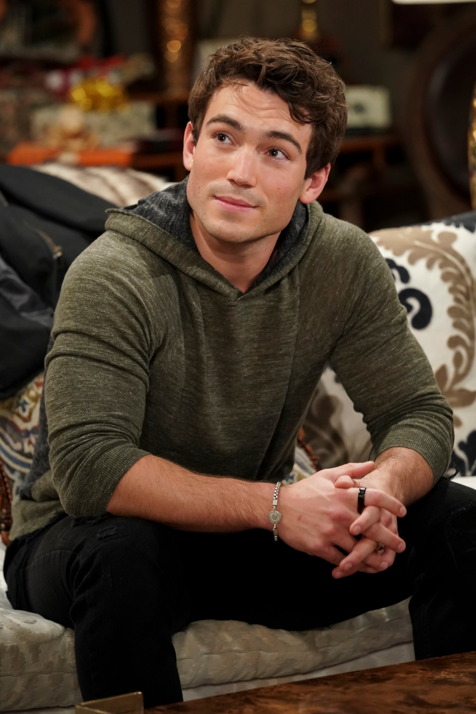 'The Young and the Restless' actor Rory Gibson wearing a grey hoodie and black pants; sits on a couch.