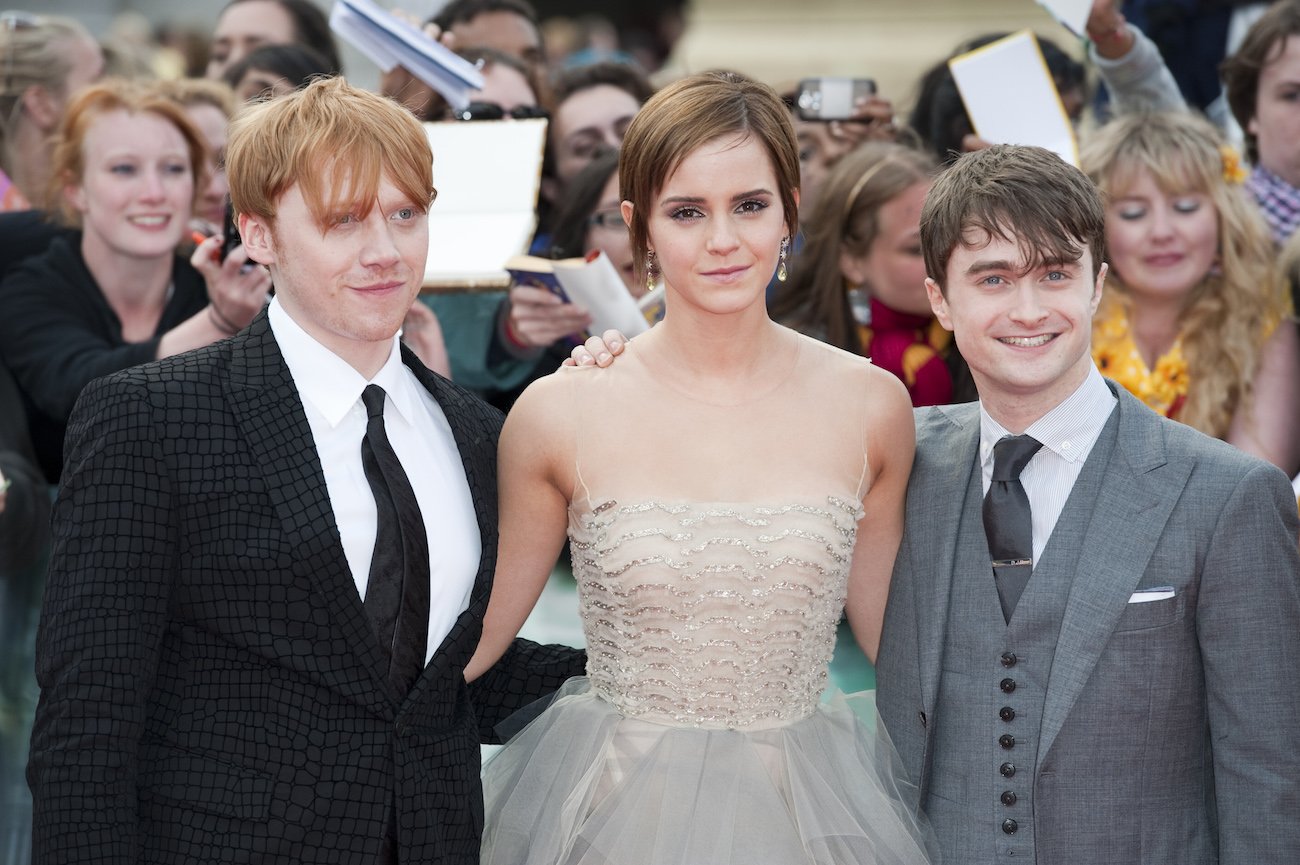Rupert Grint, Emma Watson, and Daniel Radcliffe stand together at the UK premiere of 'Harry Potter and the Deathly Hallows: Part 2.' Grint wears a black suit and tie, Watson wears a white dress, and Radcliffe wears a grey suit.