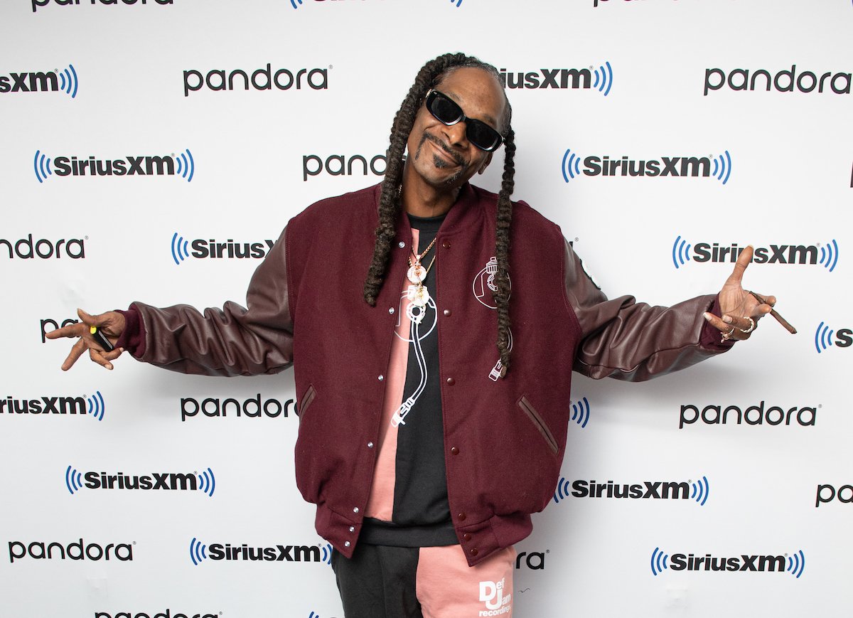 Snoop Dogg shrugging and smiling in front of a white background, holding a blunt and a lighter