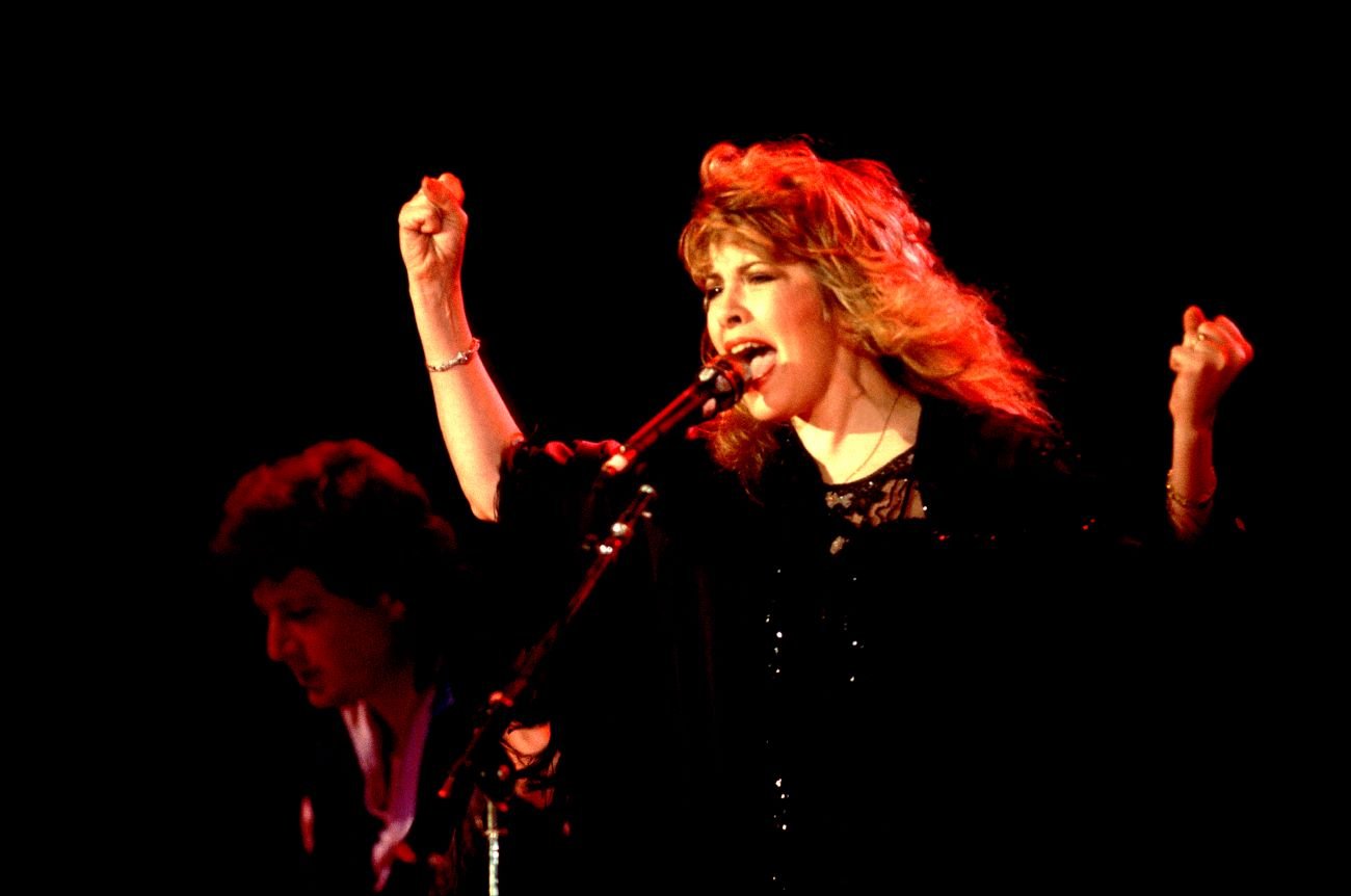 Stevie Nicks wears a black dress and sings into a microphone.