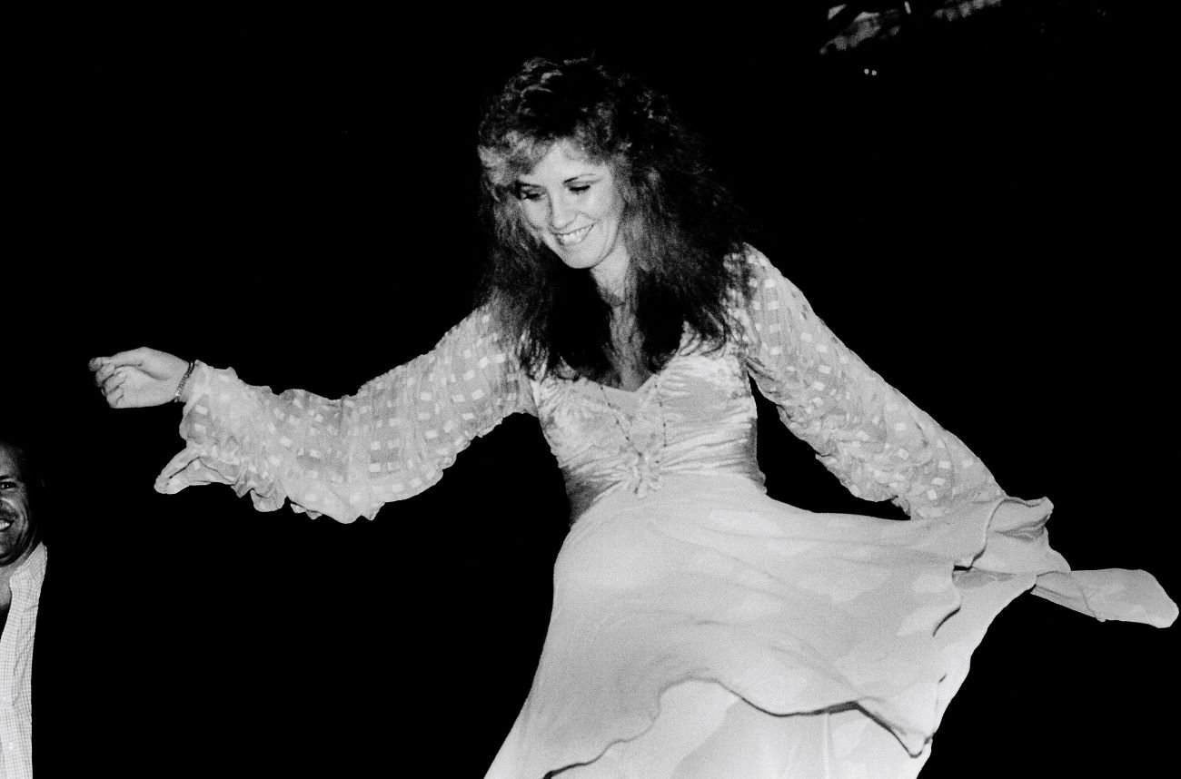 A black and white photo of Stevie Nicks wearing a long-sleeved dress and smiling.