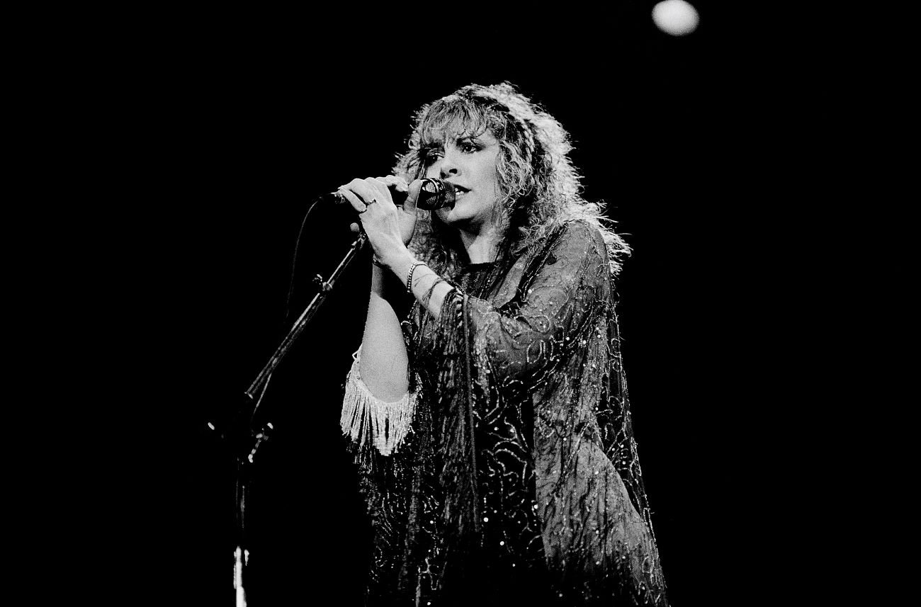 Stevie Nicks wears a beaded dress and holds a microphone while singing.