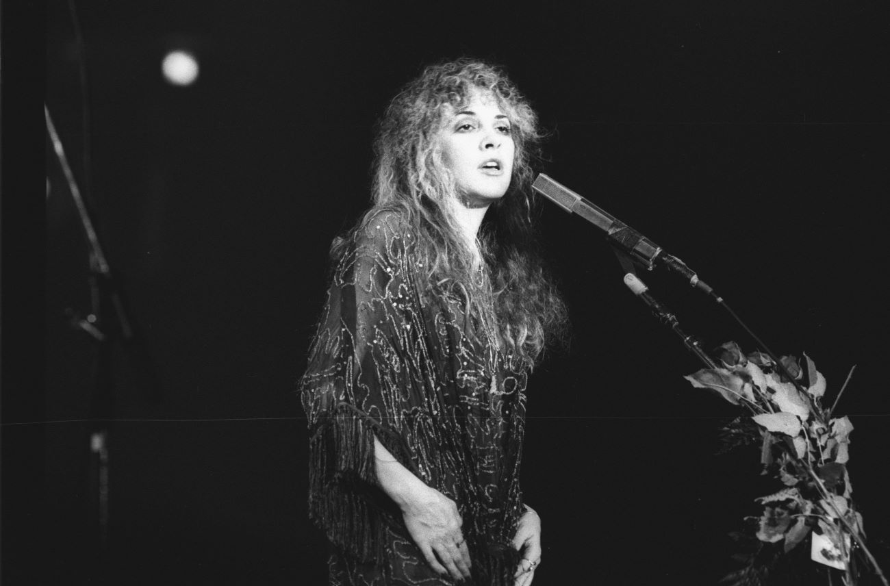 A black and white photo of Stevie Nicks wearing a beaded dress and singing into a microphone in a performance with Fleetwood Mac.