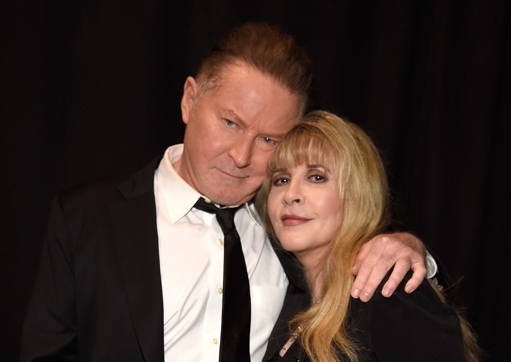 Don Henley wears a suit and holds his arm around Stevie Nicks' shoulder. She wears a black dress.
