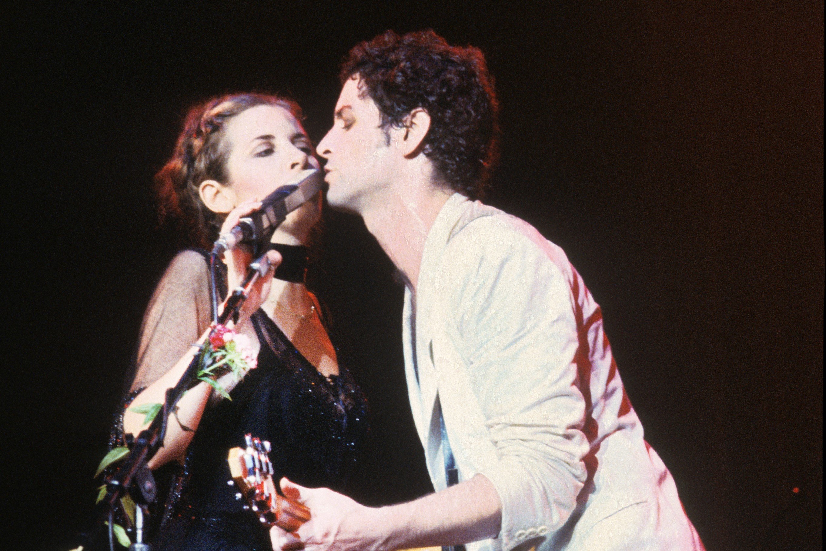 Stevie Nicks wears a black dress and sings into the same microphone as Lindsey Buckingham, who wears a white jacket. They play during a Fleetwood Mac concert.