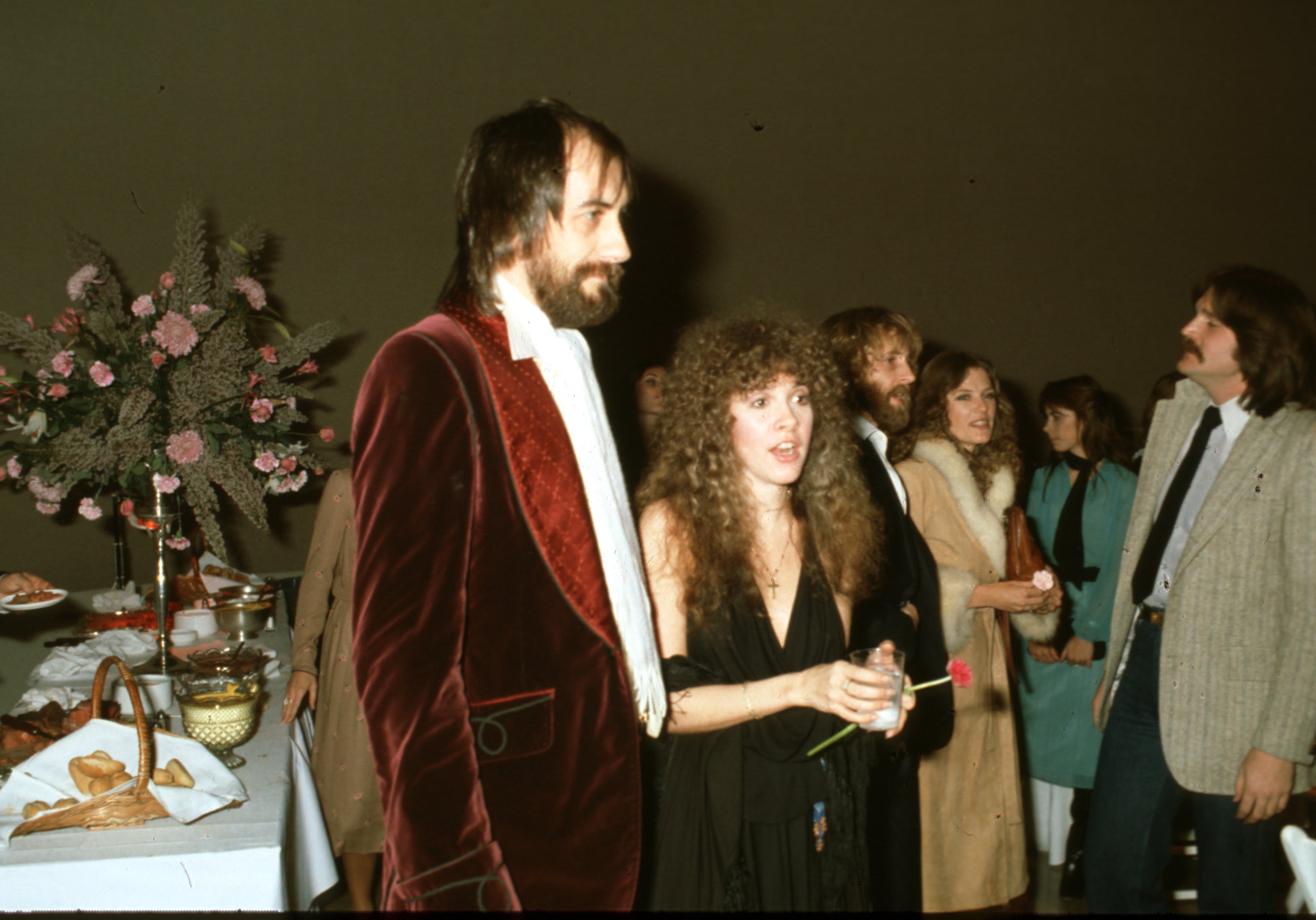 Mick Fleetwood wears a red velvet coat and Stevie Nicks wears a black dress. She holds a glass and a flower.