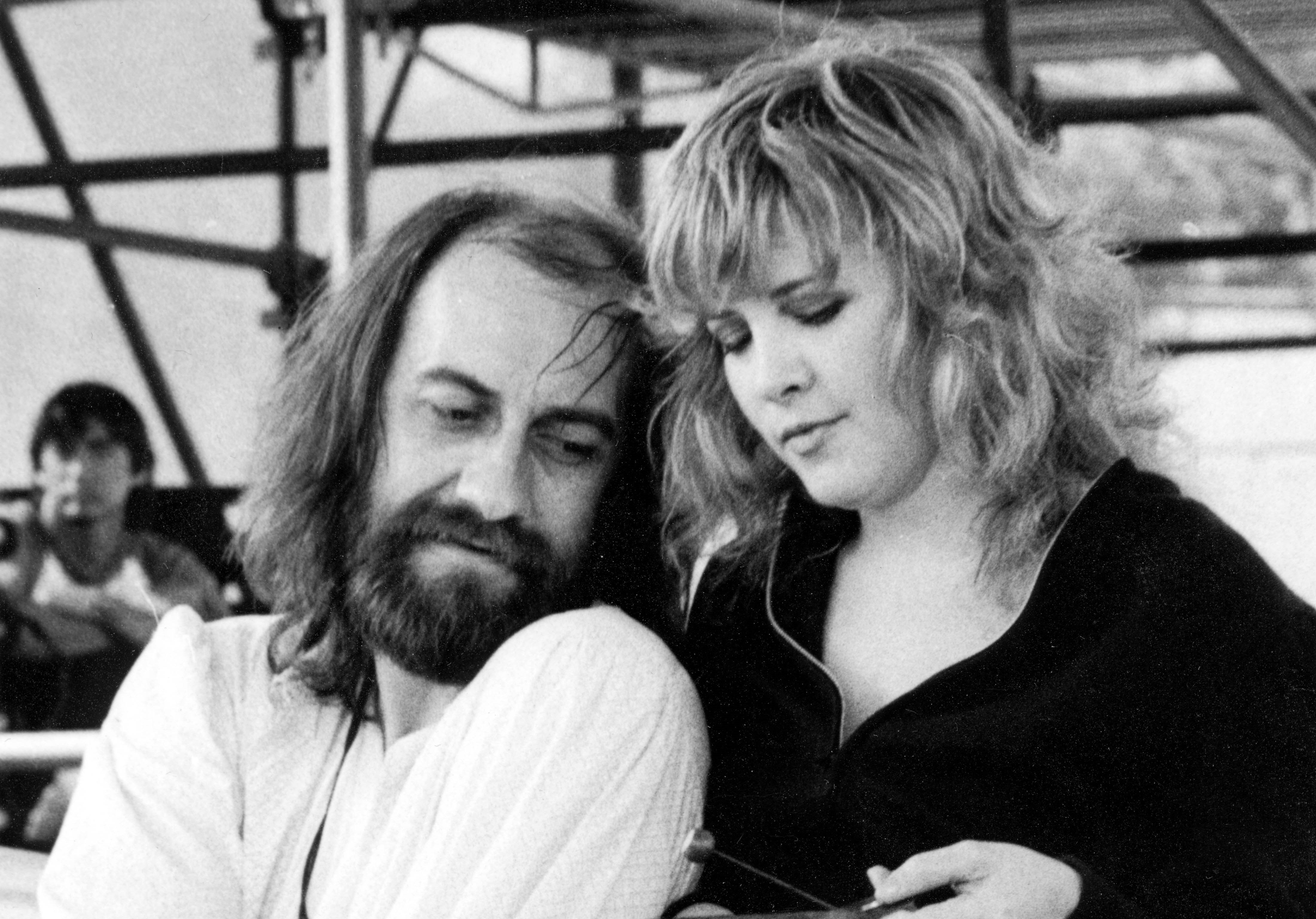 Mick Fleetwood wears a white shirt and Stevie Nicks wears a black shirt. He sits and she stands at his shoulder.