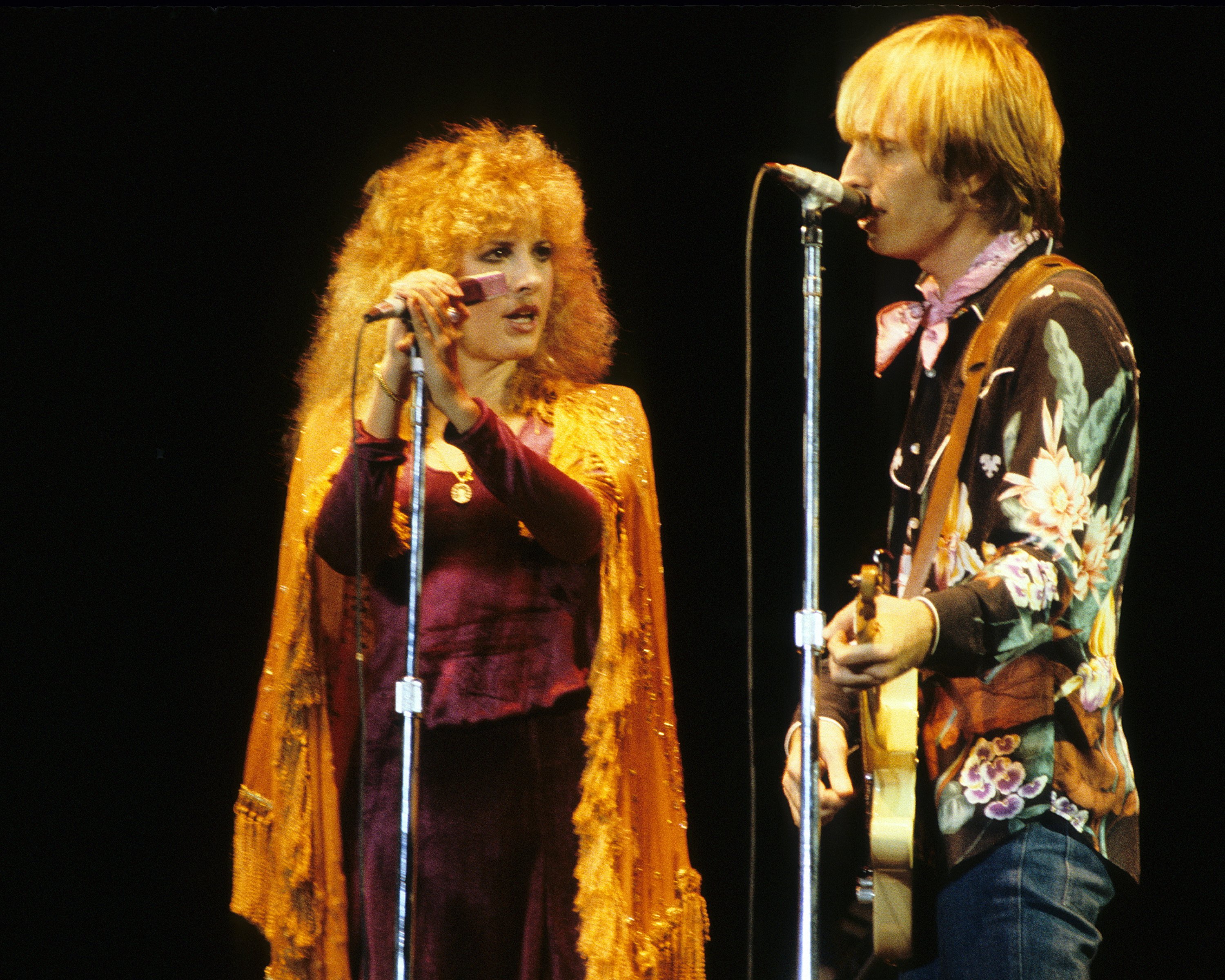 Stevie Nicks wears a shawl and holds a microphone. Tom Petty wears a floral shirt and plays guitar and sings into a microphone. 