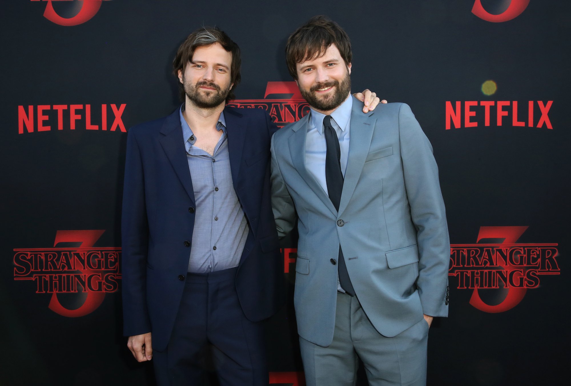 Ross and Matt Duffer, creators of 'Stranger Things,' stand in front of a 'Stranger Things 3' step and repeat while wearing suits.