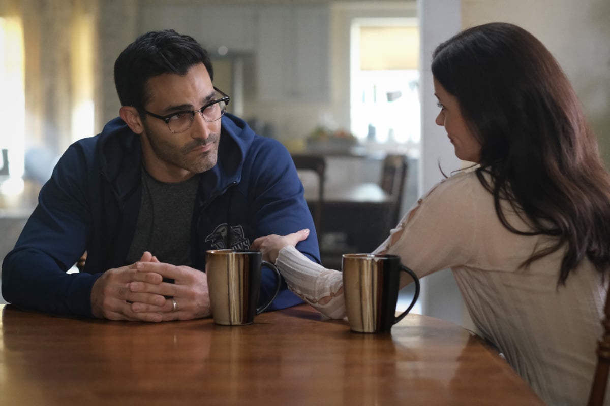 'Superman & Lois' Season 2 stars Tyler Hoechlin and Bitsie Tulloch, in character as Clark Kent and Lois Lane, talk at the kitchen table. Clark wears a blue hoodie over a gray shirt. Lois wears a white long-sleeved shirt, and she grips Clark's arm.