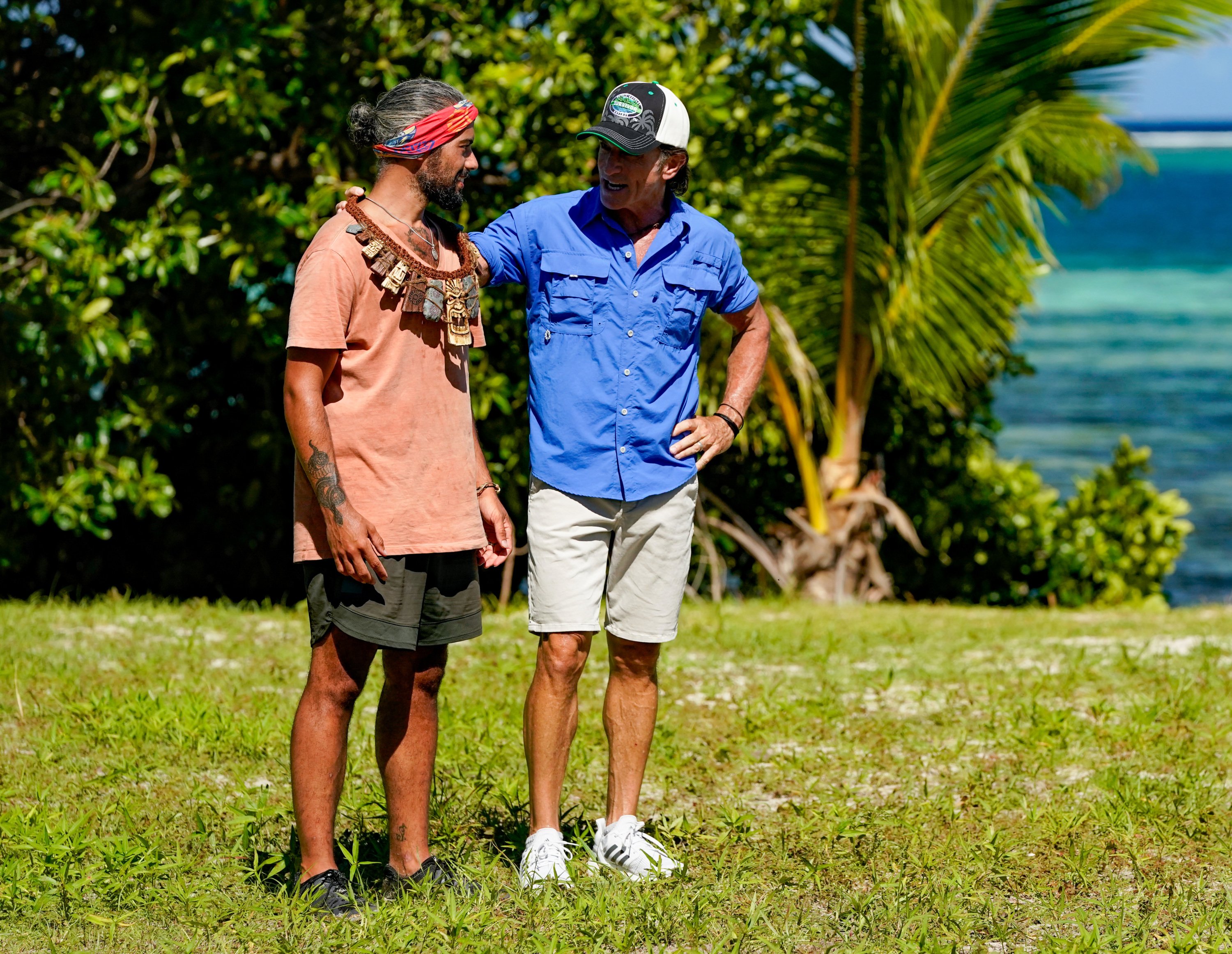 'Survivor' host Jeff Probst awards the immunity necklace to season 41 castaway Ricard Foye. Probst wears a blue button-up shirt, tan shorts, and a 'Survivor' baseball cap. Ricard wears a salmon-colored shirt, green shorts, and his 'Survivor' buff around his head.