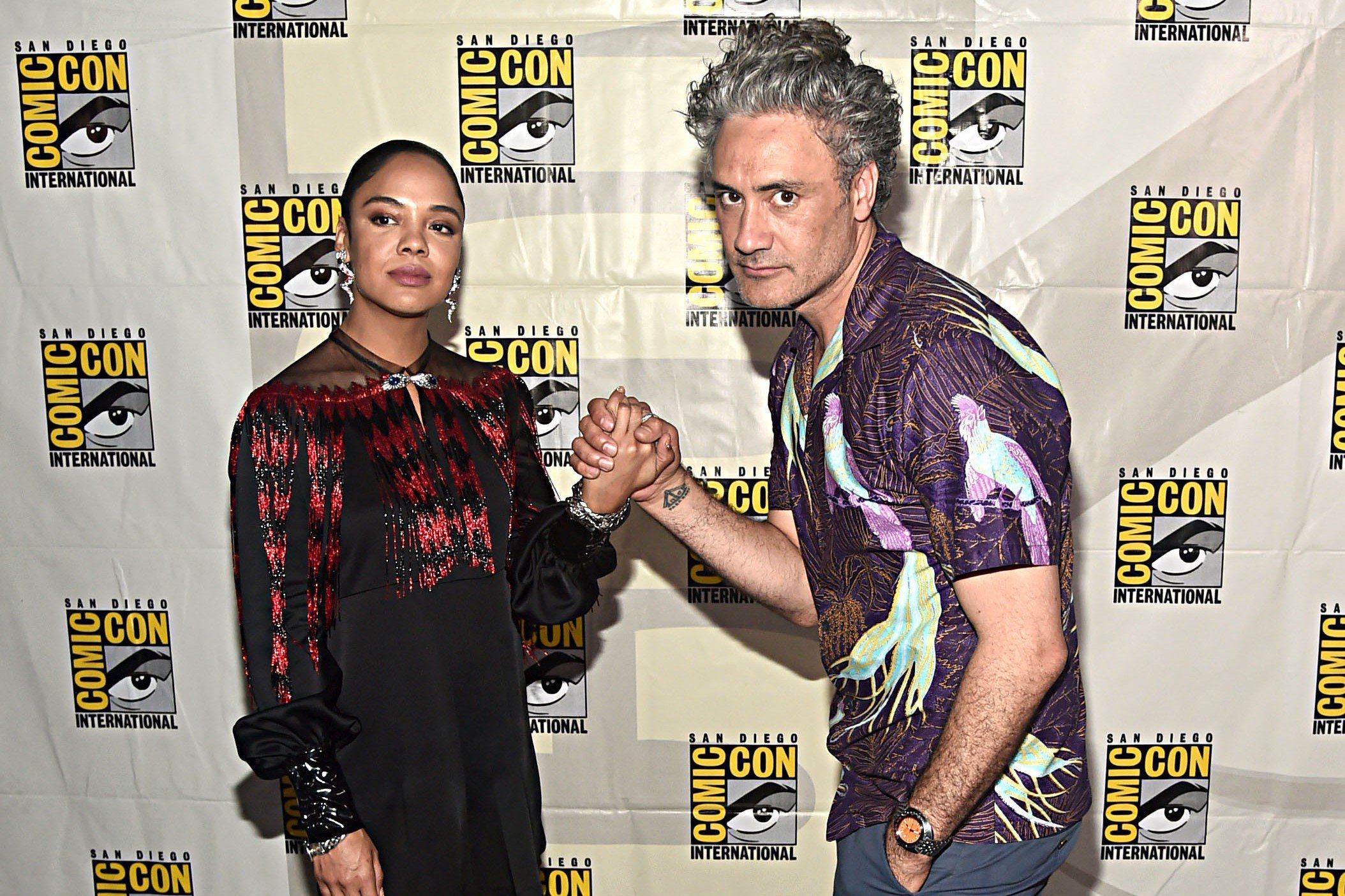 'Thor: Love and Thunder' star Tessa Thompson and director Taika Waititi hold hands while posing for pictures. Thompson wears a red and black long-sleeved dress. Waititi wears a purple, gold, and blue shirt with birds and tropical patterns.