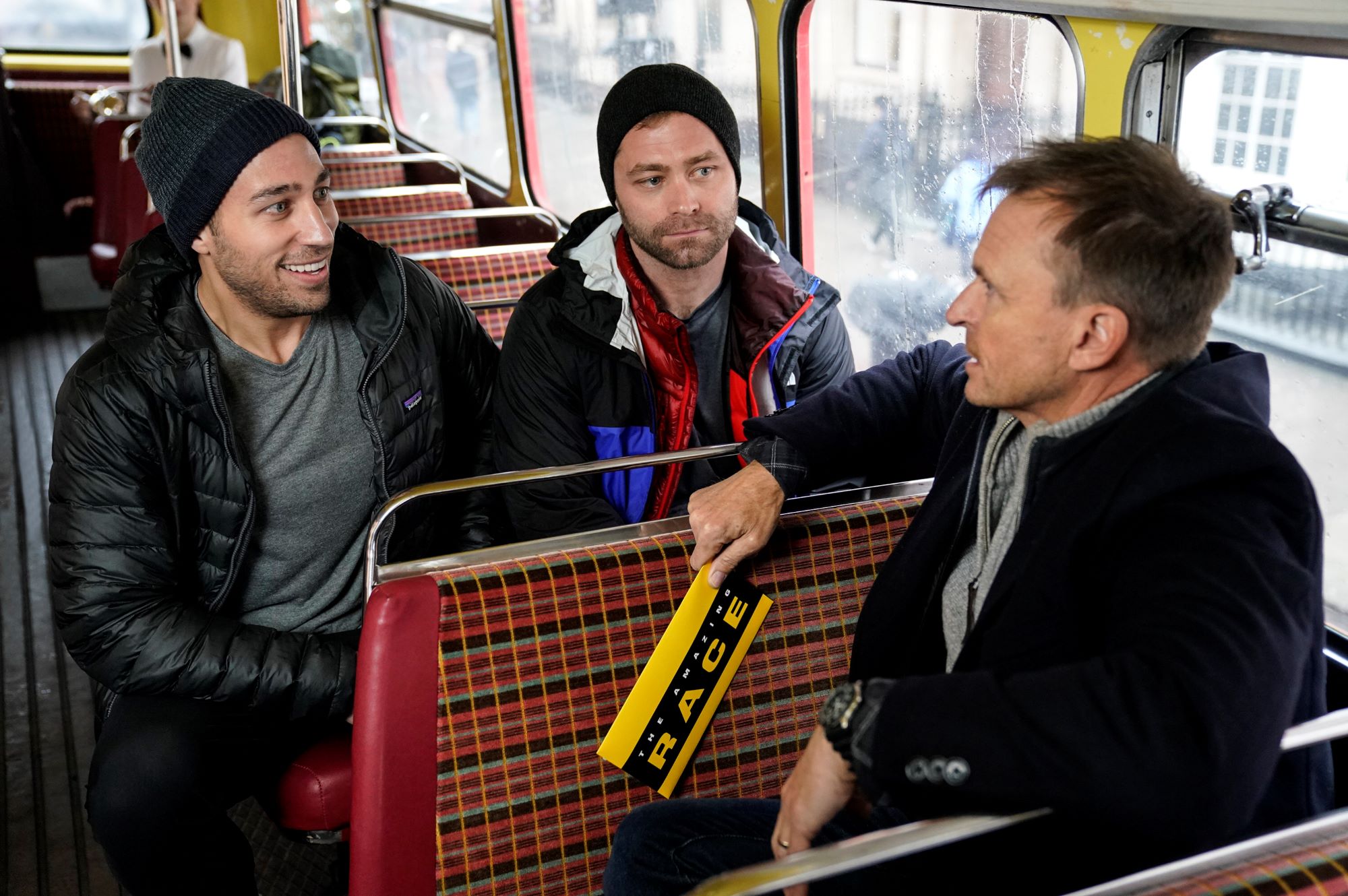 'The Amazing Race' Season 33 stars Ryan Ferguson, Dusty Harris, and Phil Keoghan talk in a double-decker bus during a pit stop in London. Ferguson wears a black jacket over a gray shirt and black beanie. Harris wears a black, blue, and red jacket and a black beanie. Keoghan wears a black coat over a gray sweater.