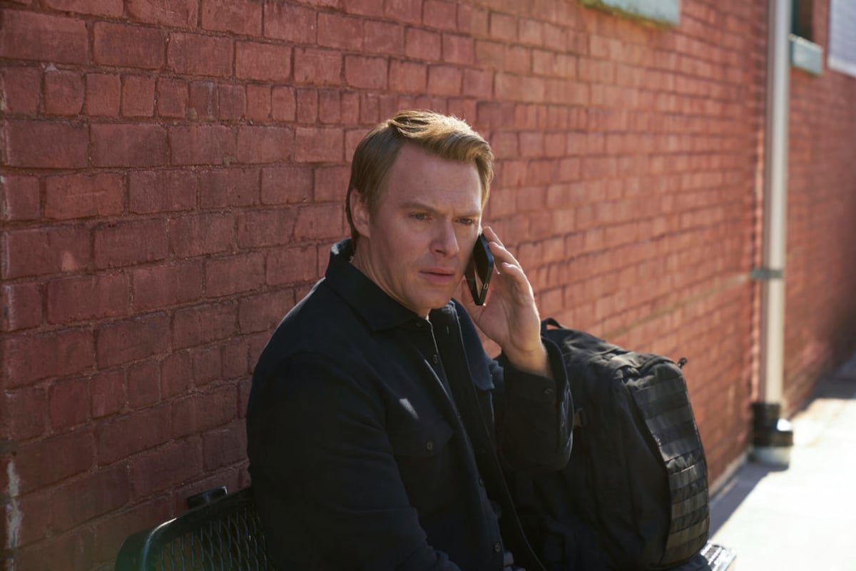 Diego Klattenhoff as Donald Ressler in The Blacklist Season 9. Ressler sits in front of a brick wall and talks on his cellphone. 