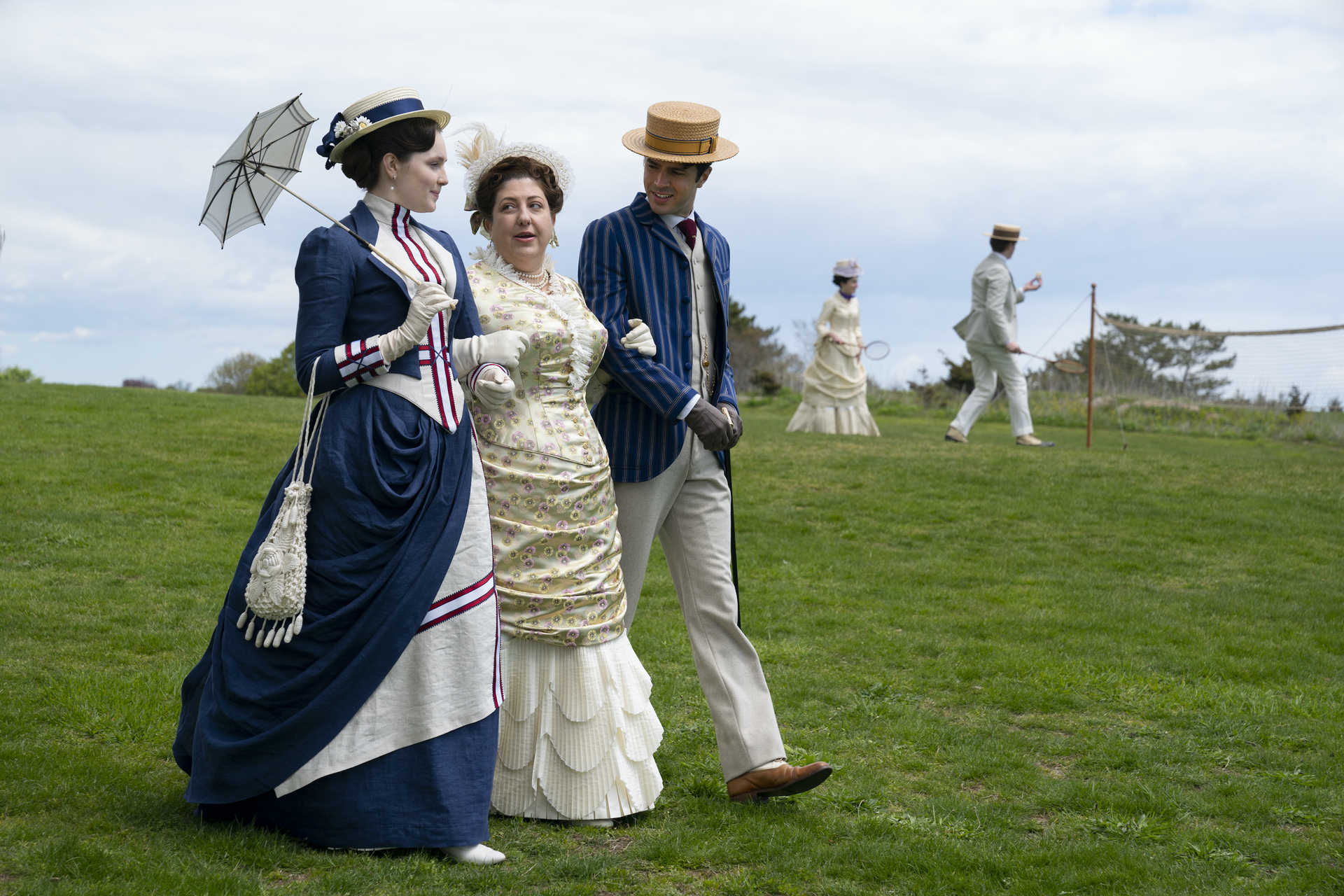 Two women, one holding a parasol, and a man in a straw boater hat, walking across a lawn in 'The Gilded Age' 