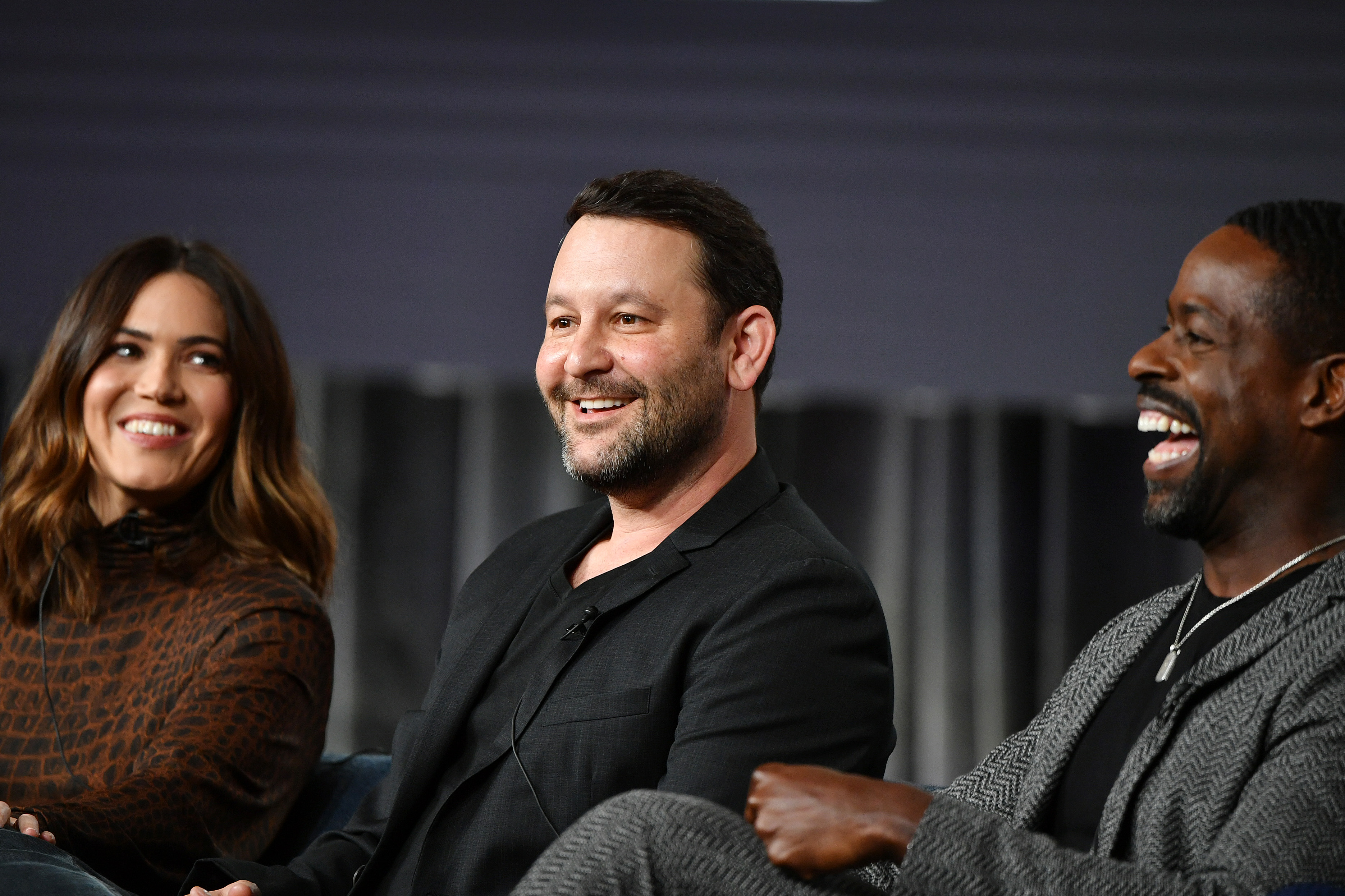 'This Is Us' Season 6 cast and crew, Mandy Moore, Dan Fogelman, and Sterling K. Brown, gather onstage for an event. Moore wears a brown and black sweater. Fogelman wears a black blazer over a black shirt. Brown wears a gray blazer over a black shirt. The 'This Is Us' series finale hasn't been written yet.