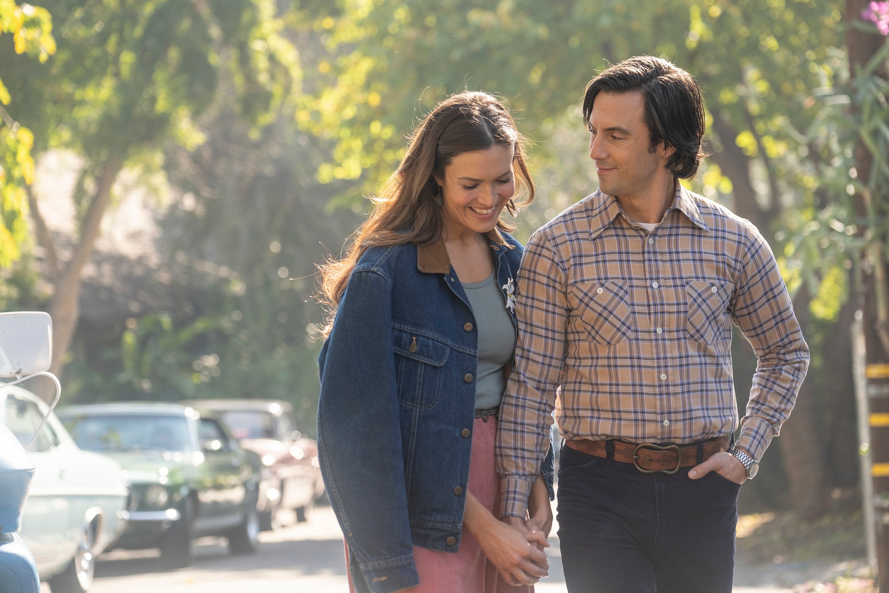 'This Is Us' stars Mandy Moore and Milo Ventimiglia, in character as Rebecca and Jack Pearson, walk hand-in-hand down a street. Rebecca wears a jean jacket over a green top and pink bottoms. Jack wears a yellow and blue plaid shirt and jeans.