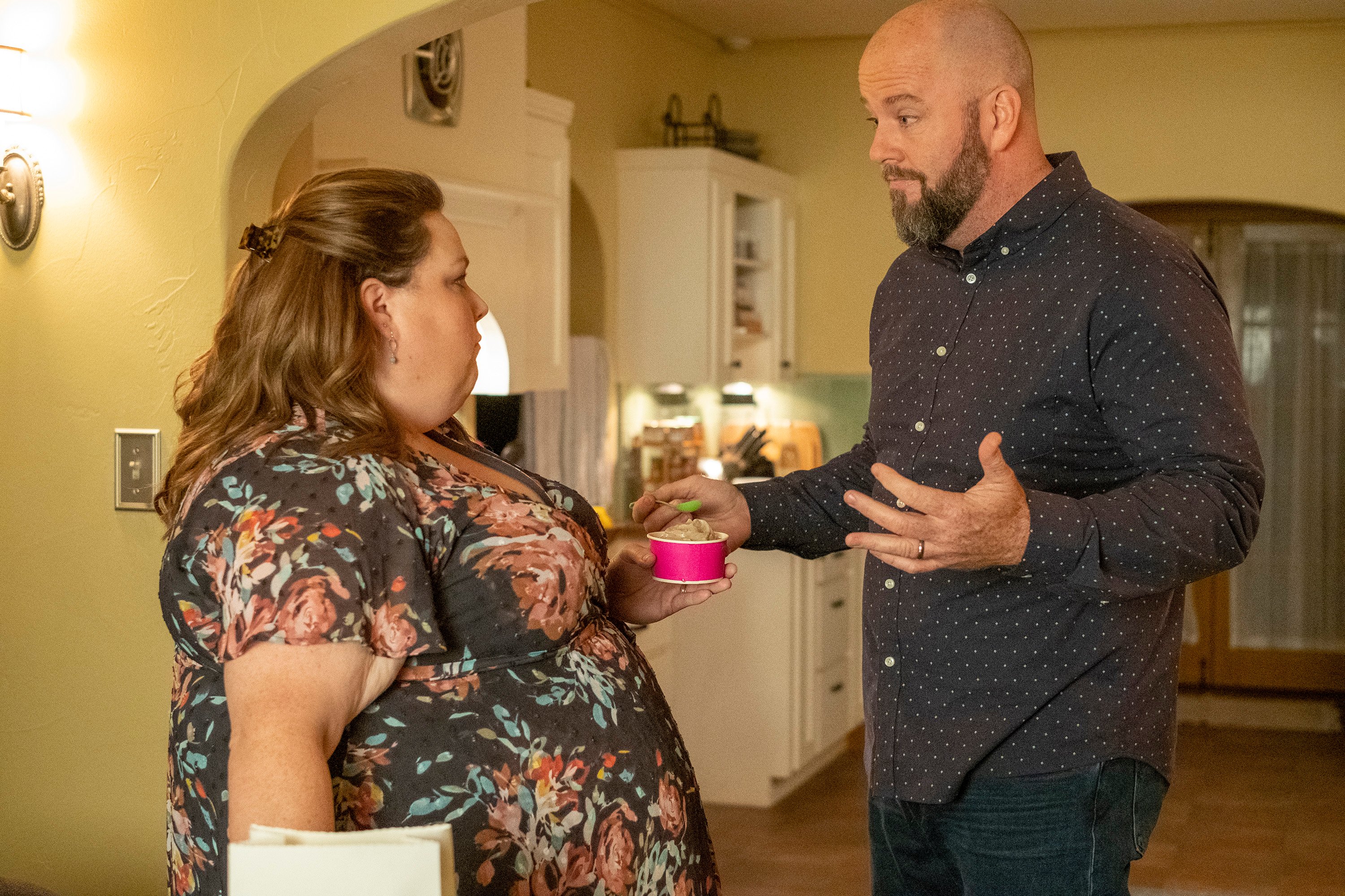 'This Is Us' Season 6 Episode 3 stars Chrissy Metz and Chris Sullivan, in character as Kate and Toby, talk in their kitchen. Kate wears a blue and pink floral dress and holds a cup of ice cream. Toby wears a dark blue button-down shirt with white polka dots and jeans.