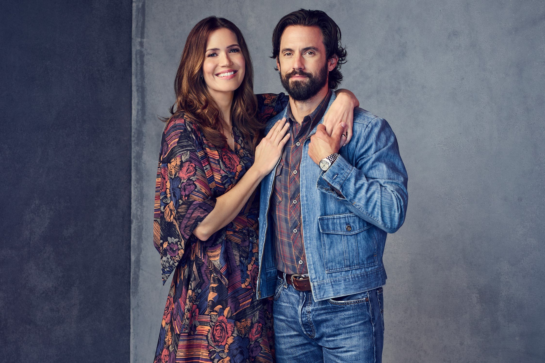 'This Is Us' Season 6 actors Mandy Moore and Milo Ventimiglia, in character as Rebecca and Jack Pearson, pose for promotional pictures. Moore wears a multi-colored floral patterned dress. Ventimiglia wears a jean jacket over a red, blue, and yellow plaid button-up shirt and jeans.
