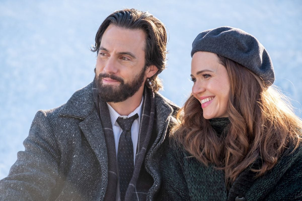 'This Is Us' Season 6 Episode 4 actors Milo Ventimiglia and Mandy Moore, in character as Jack and Rebecca Pearson, share a scene outside. Jack wears a gray coat over his black suit and black tie. Rebecca wears a dark green cardigan and a gray hat.