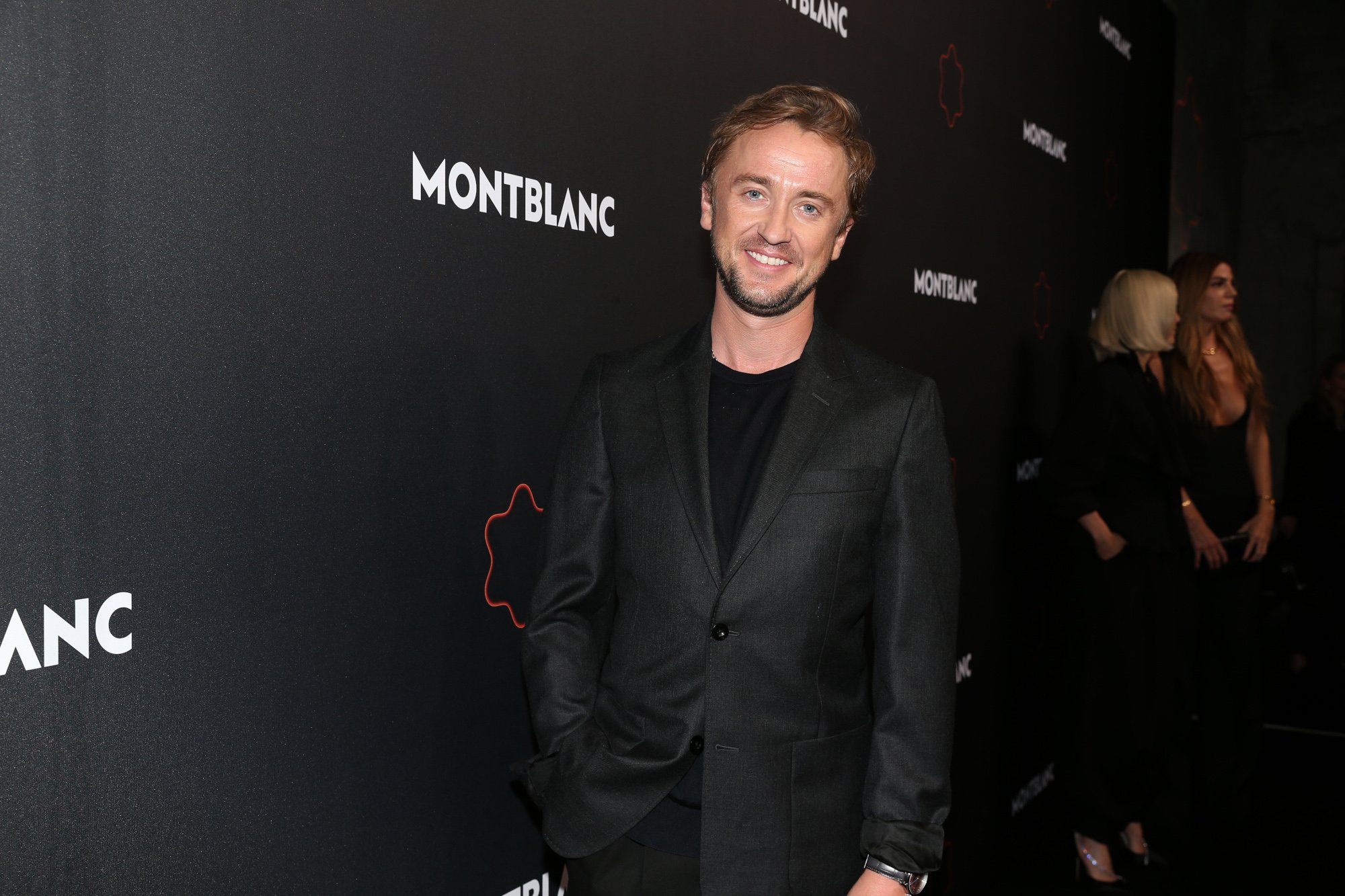 'Harry Potter' star Tom Felton wearing an all-black suit and standing in front of a Montblanc wall