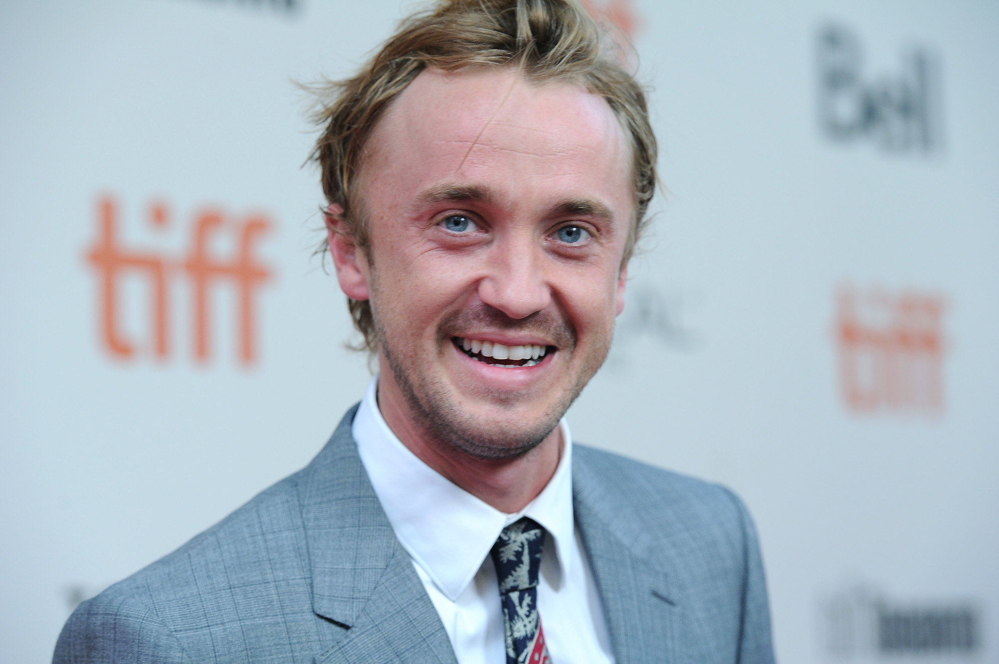 'Harry Potter' star Tom Felton wearing a gray suit and smiling. Tom Felton's net worth in 2022 is estimated at $20 million.