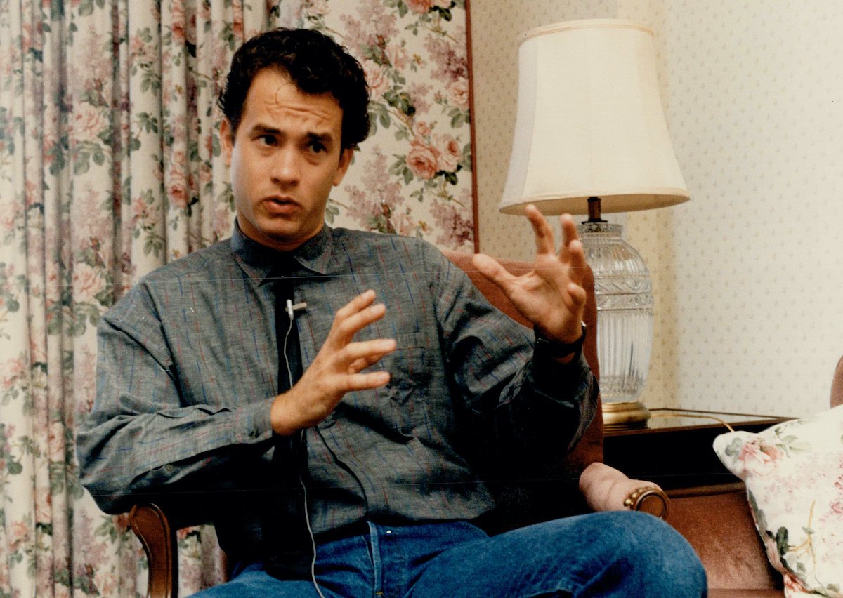 Tom Hanks wears a gray shirt and blue jeans as he speaks with his hands out