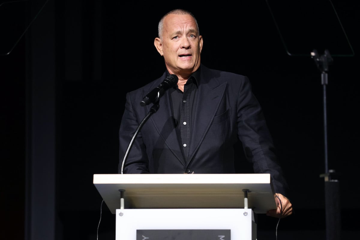Tom Hanks wears a dark suit as he speaks at a podium