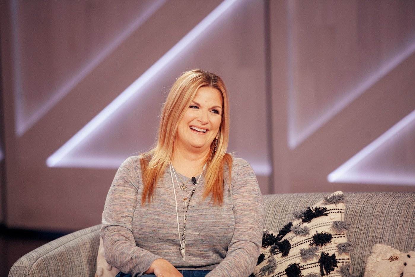 Trisha Yearwood smiles as she sits on a couch and looks on