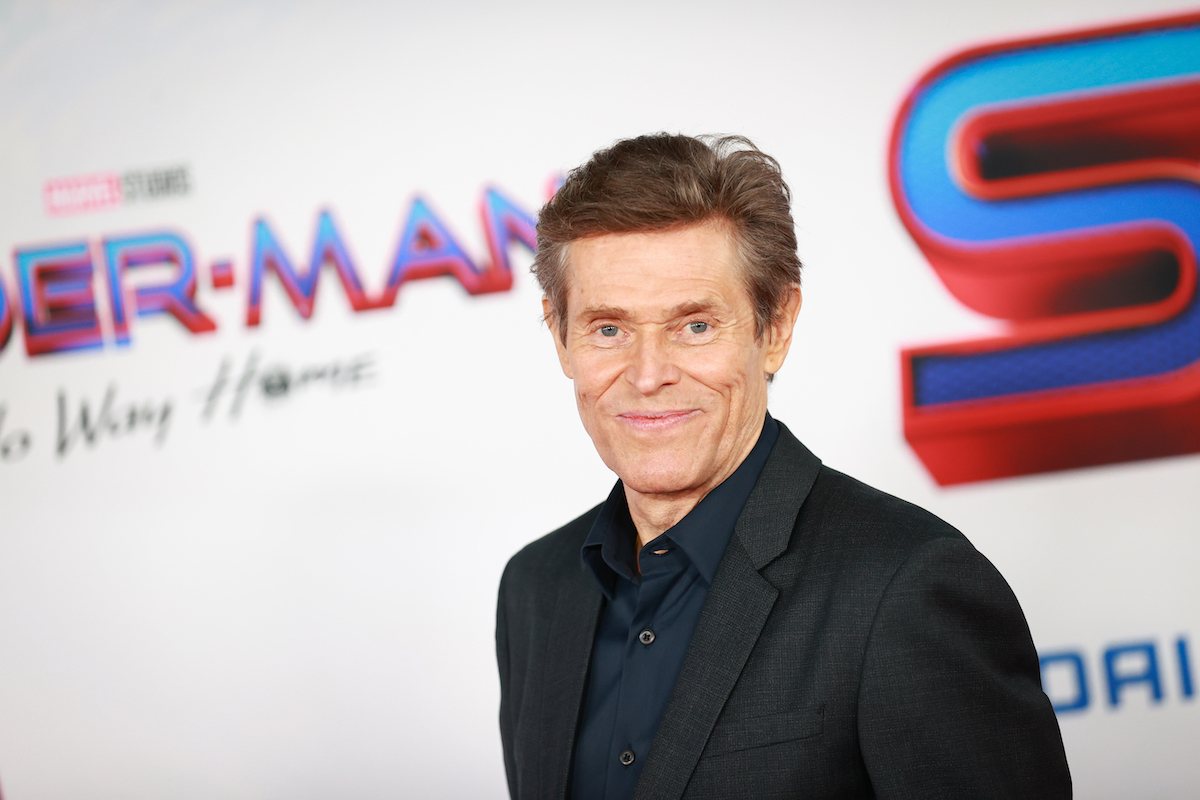 Willem Dafoe smiling in front of a white background