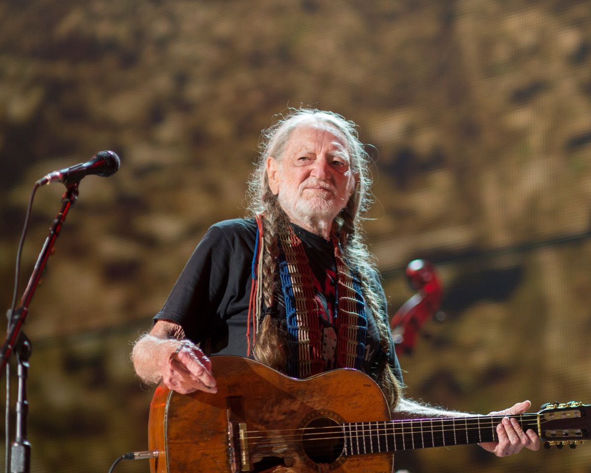 Willie Nelson plays his guitar, wearing a t-shirt with his hair in braids