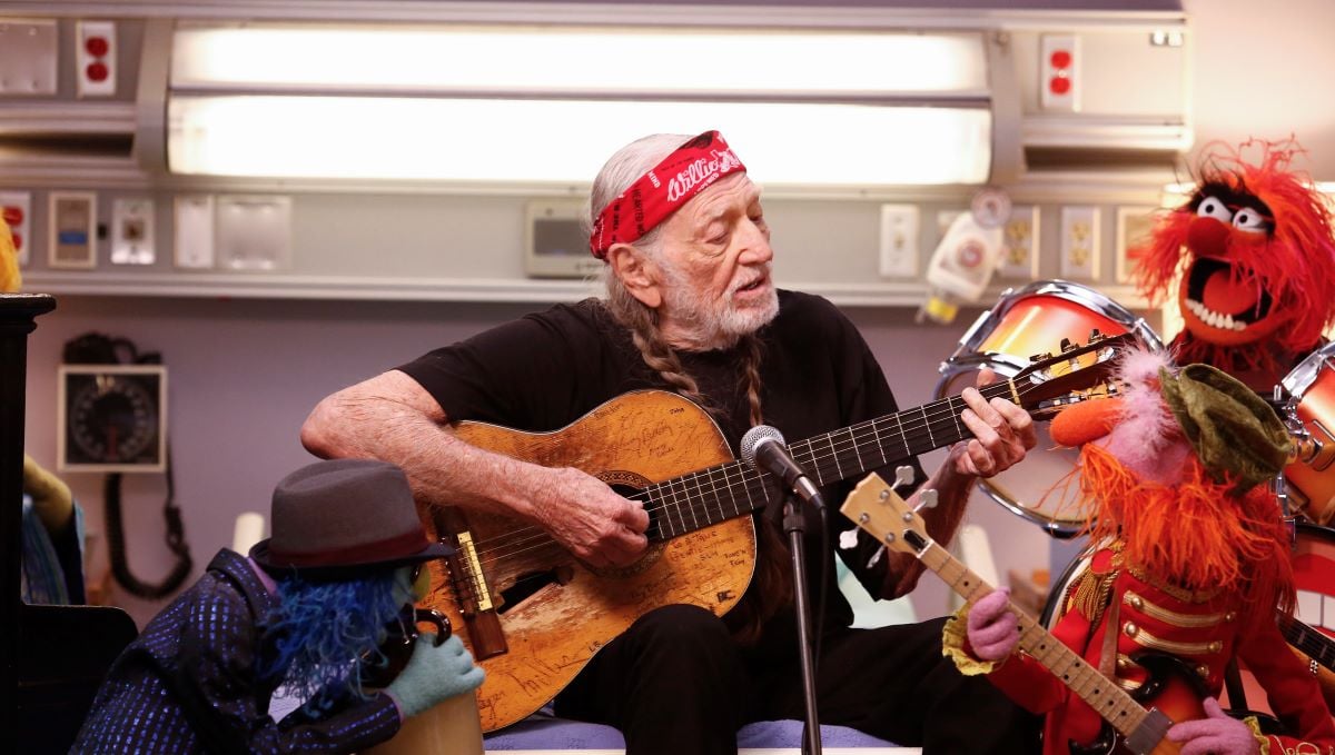 Willie Nelson in a red bandana, holding his guitar while surrounded by Muppets with instruments