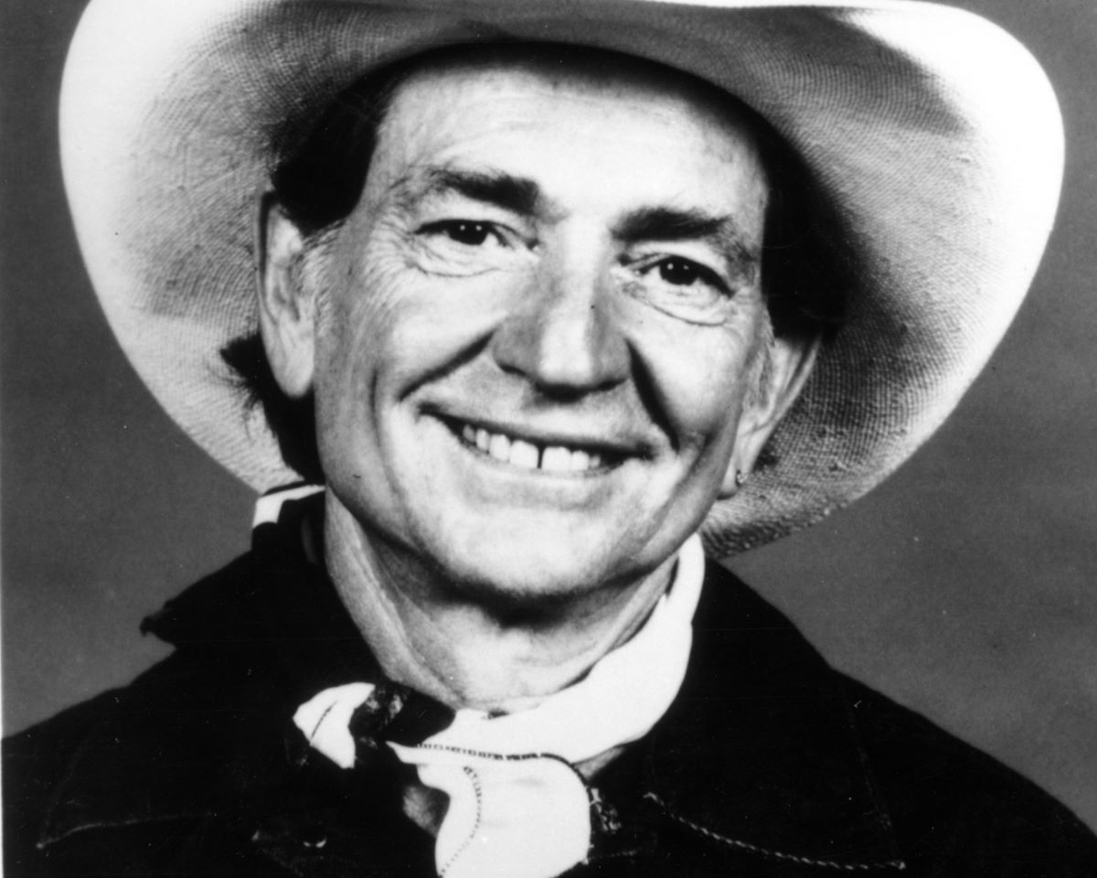 Black and white photo of Willie Nelson smiling, wearing a cowboy scarf and hat
