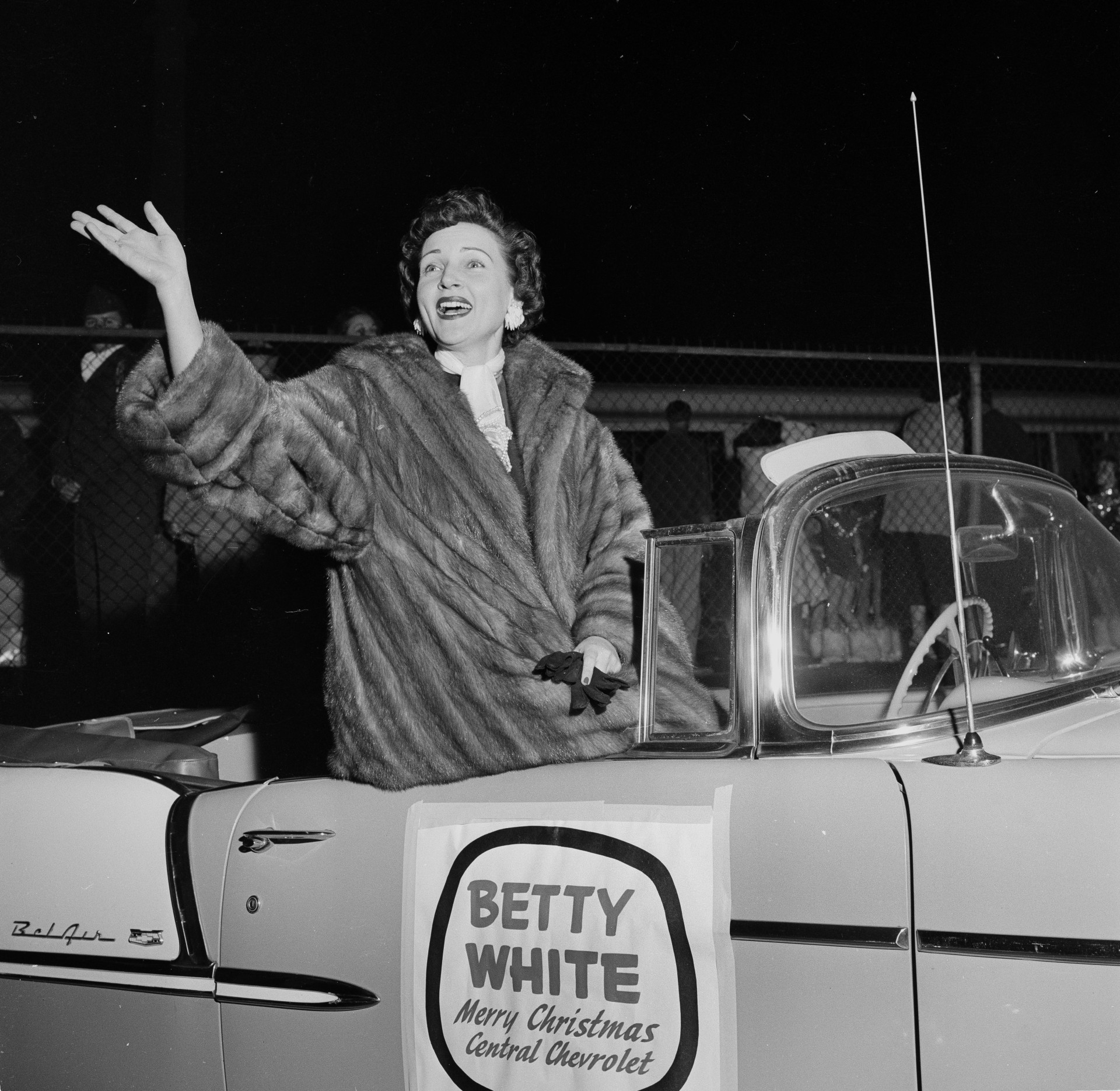 Betty White waves from a Chevrolet while wearing a fur coat