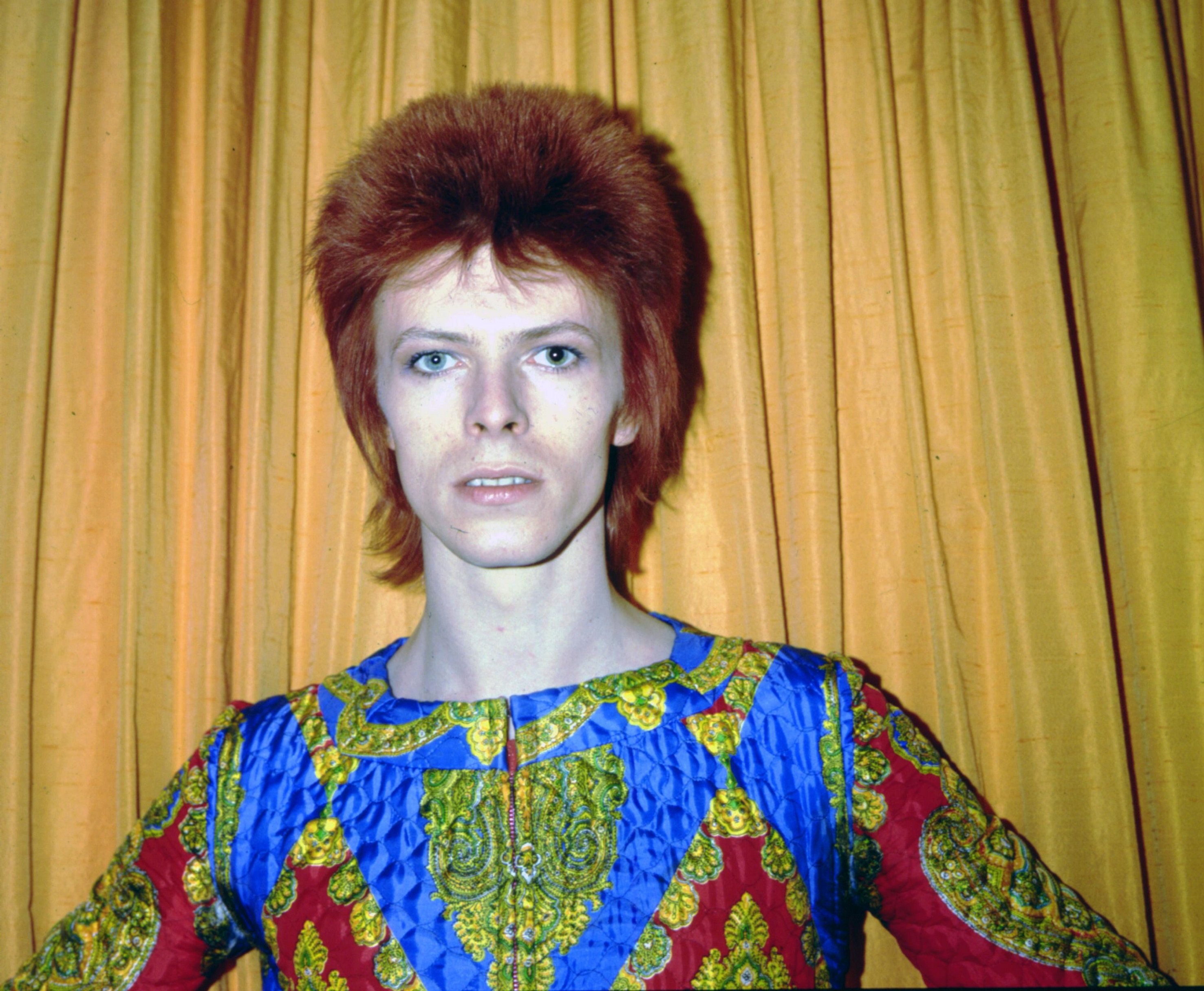 David Bowie dressed as Ziggy Stardust and standing in front of a curtain